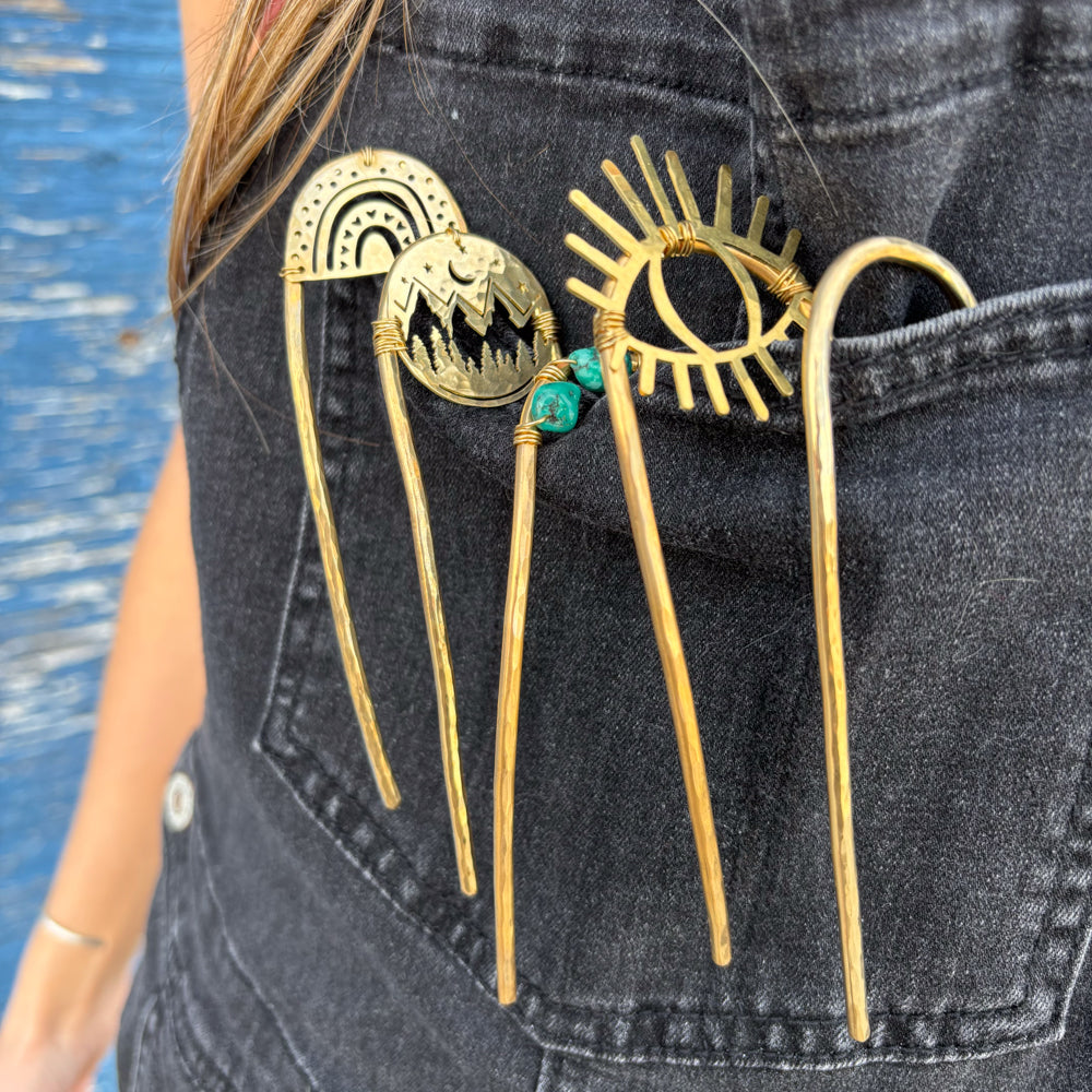 Four decorative gold hairpins with unique designs, including a rainbow, mountains, an eye, and a spiral, are tucked into the front pocket of a black denim outfit.