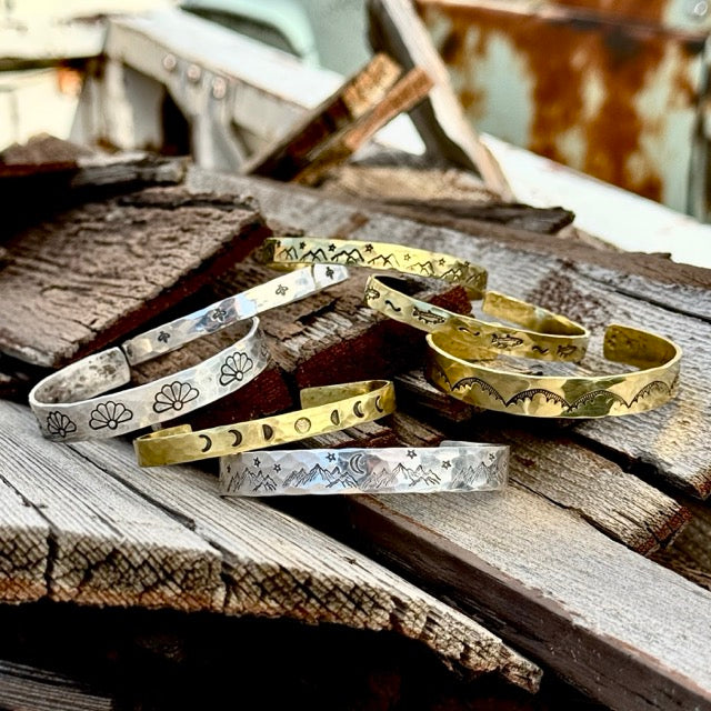 Three metal hair forks with decorative ends and two engraved metal cuff bracelets are displayed on weathered wood. The hair forks feature floral, turquoise, and mountain designs.