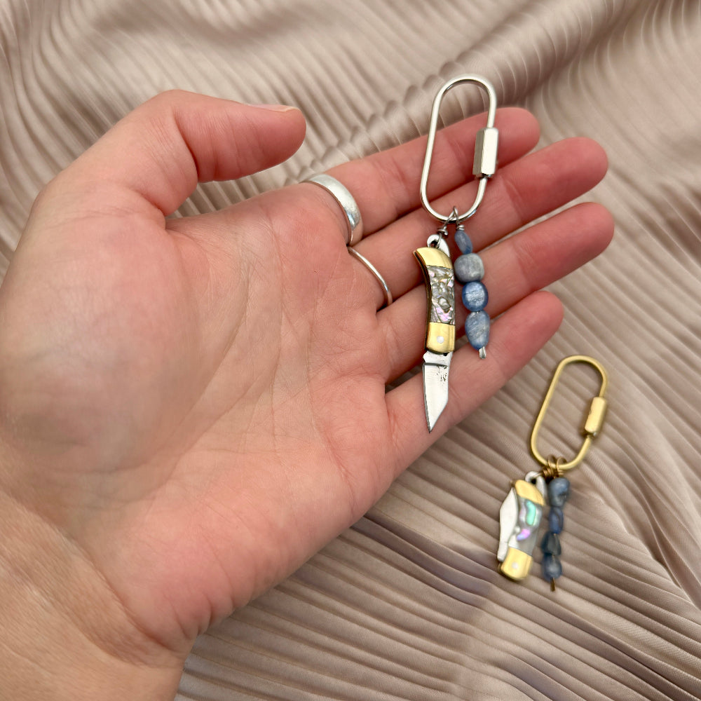 Hand holding two keychain purse chars with knife designs on a beige fabric background