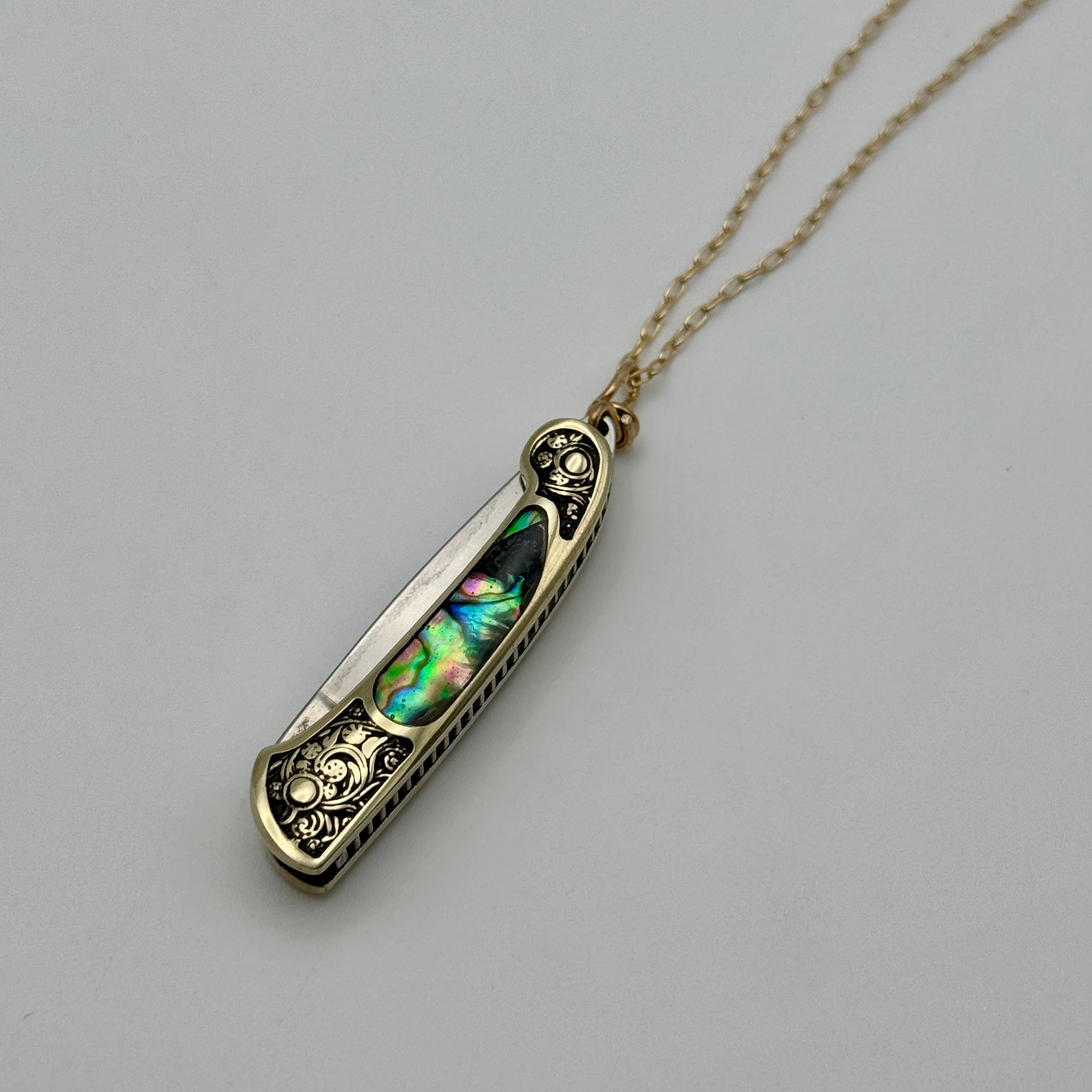 The Lennox Necklace by Fluff Hardware features a pocket knife with ornate gold detailing and an iridescent abalone inlay, suspended from a delicate gold chain and displayed on a plain light background.