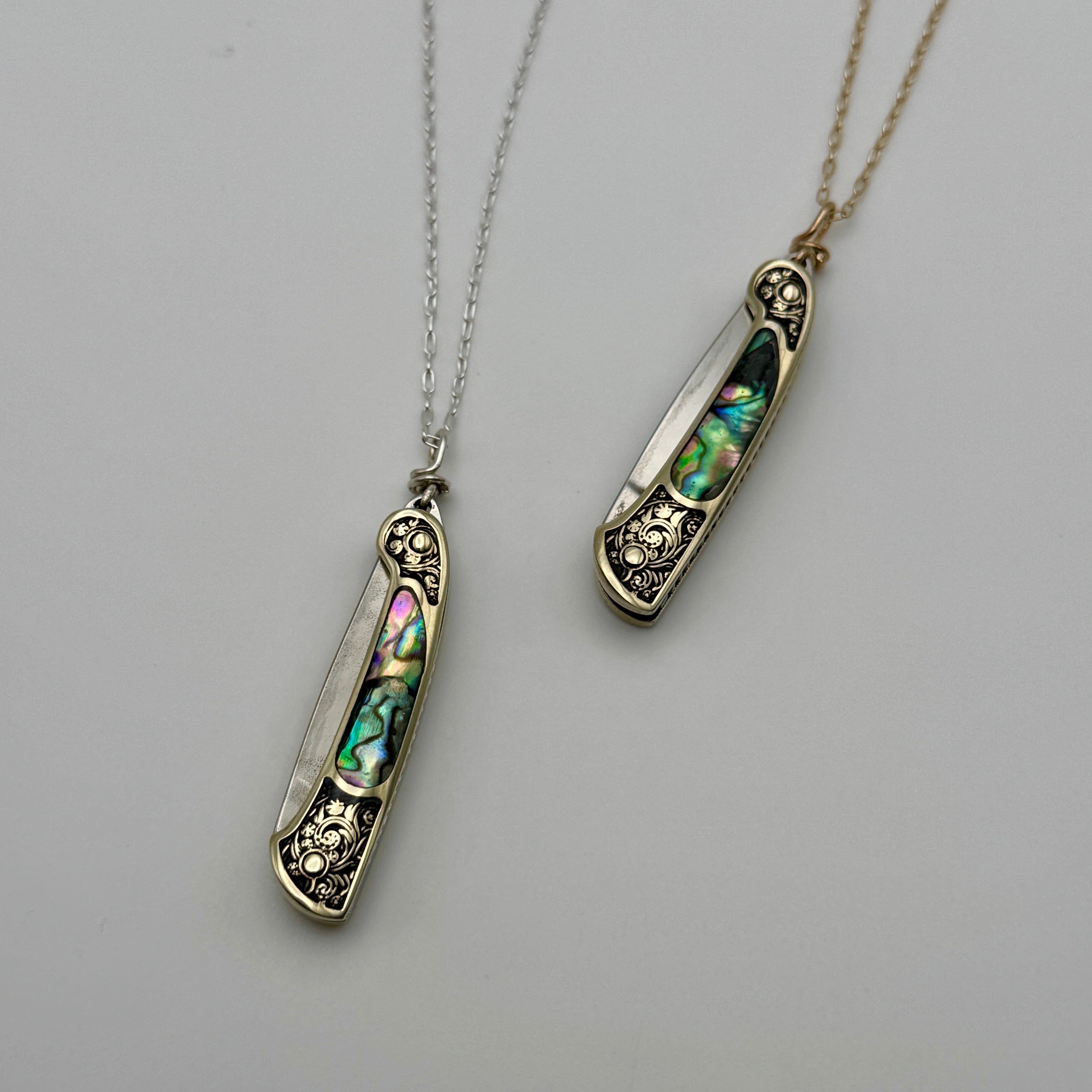 The Lennox Necklace by Fluff Hardware features two ornate pocket knife pendants with engraved details and iridescent abalone shell inlays. One necklace comes on a silver-toned chain, the other on a gold-toned, both displayed against a light background.