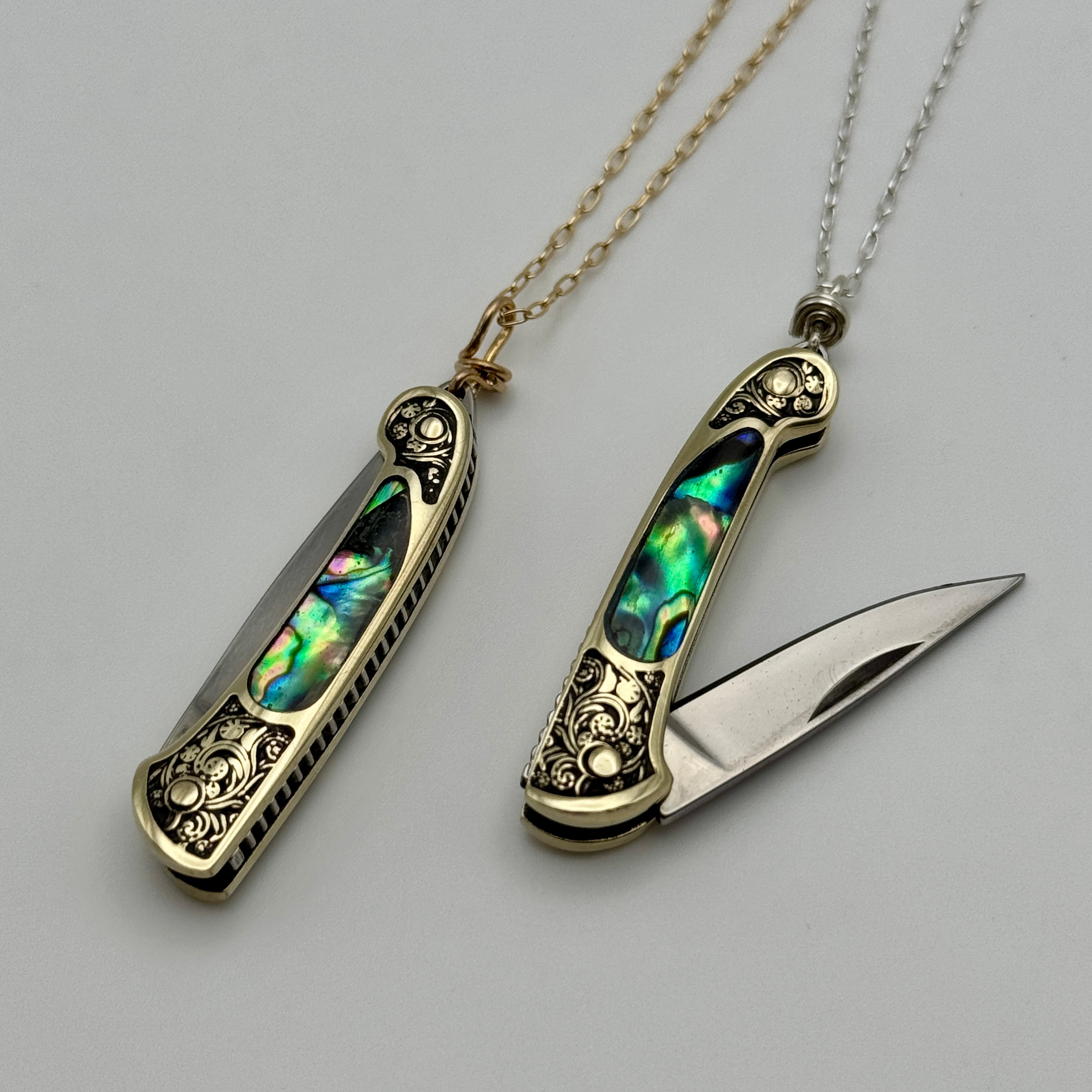 Two ornate Fluff Hardware Lennox Necklace pocket knives with colorful abalone inlays are displayed as functional jewelry—one closed on a gold chain, the other open on a silver chain—both set against a plain light background.