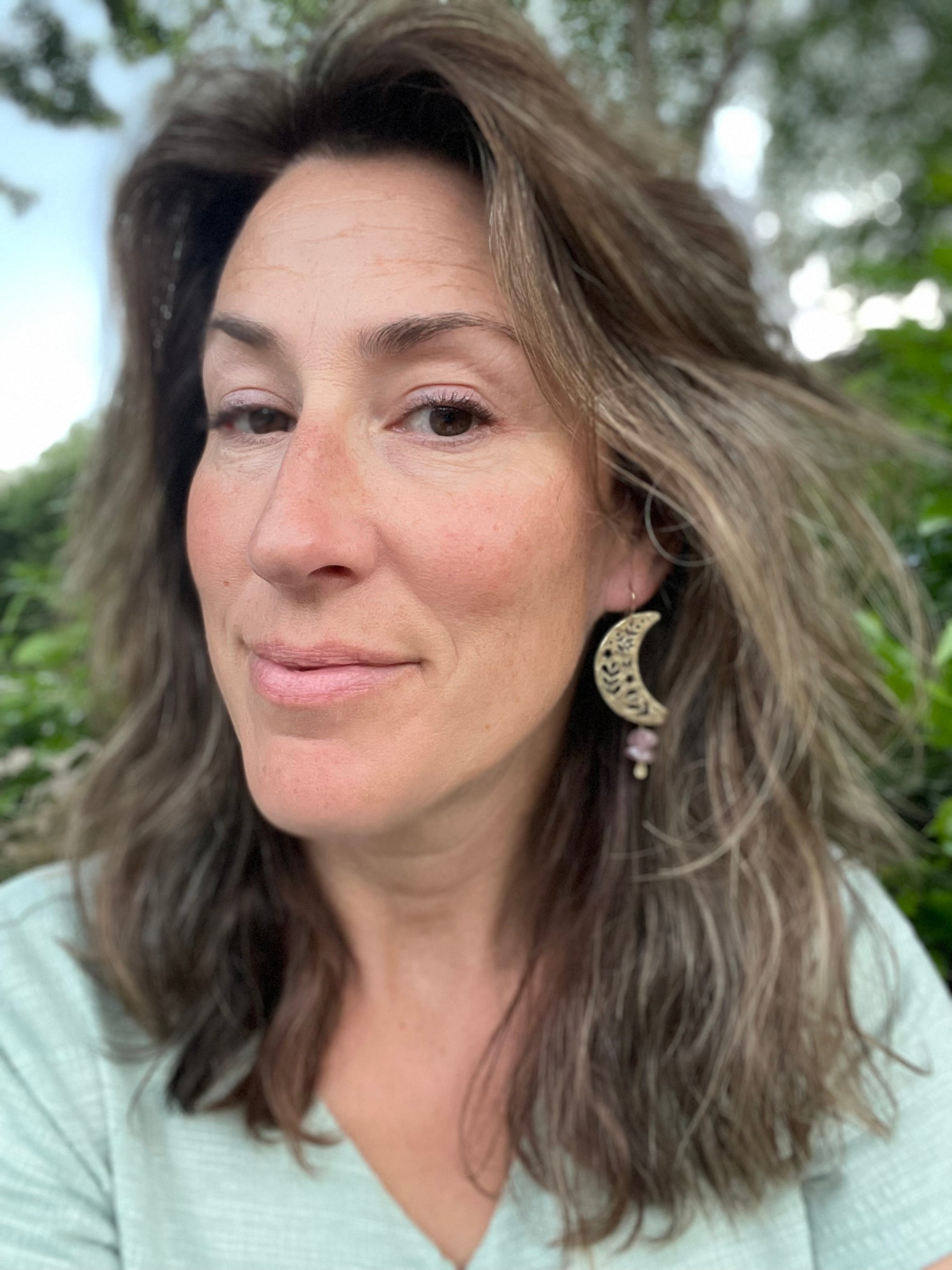 A woman with wavy brown hair and subtle highlights wears a light green top and Fluff Hardware’s Moon Garden Earrings, standing outdoors with softly blurred greenery and trees in the background.