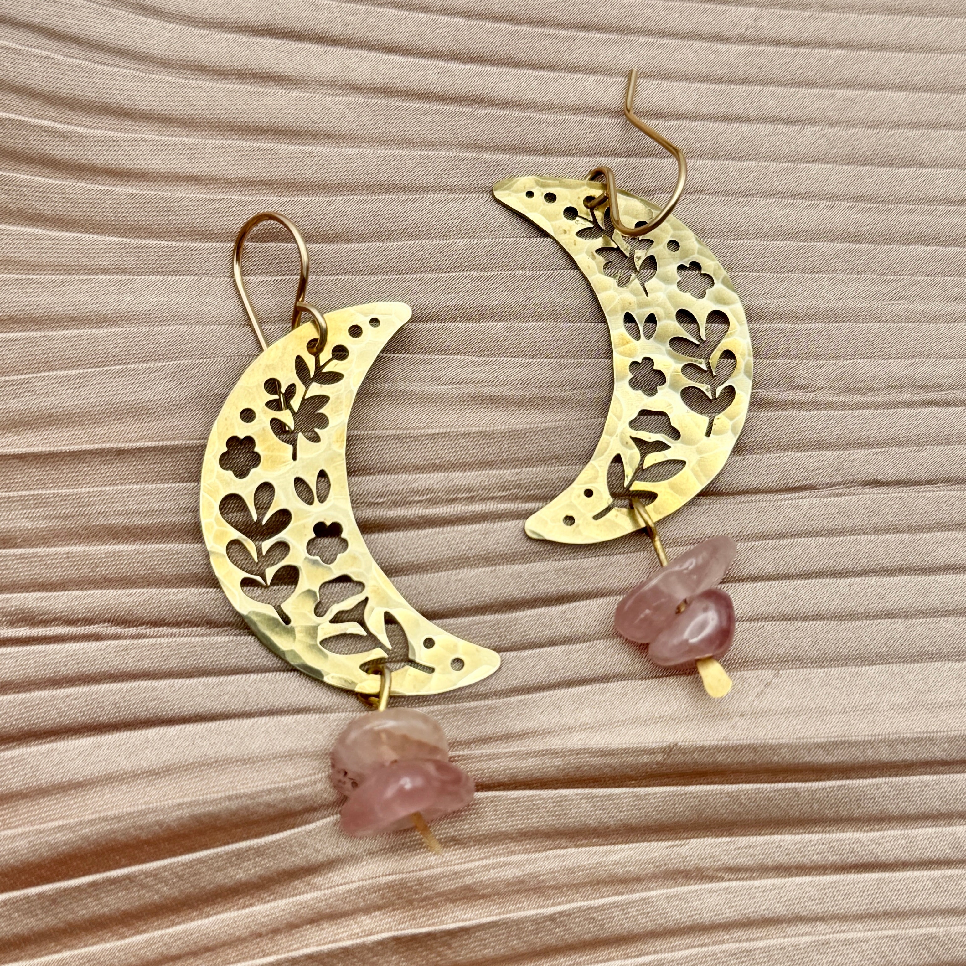 The Moon Garden Earrings by Fluff Hardware feature crescent-shaped gold designs with floral cutouts and dangling rose quartz beads, showcased on a textured light beige fabric background.