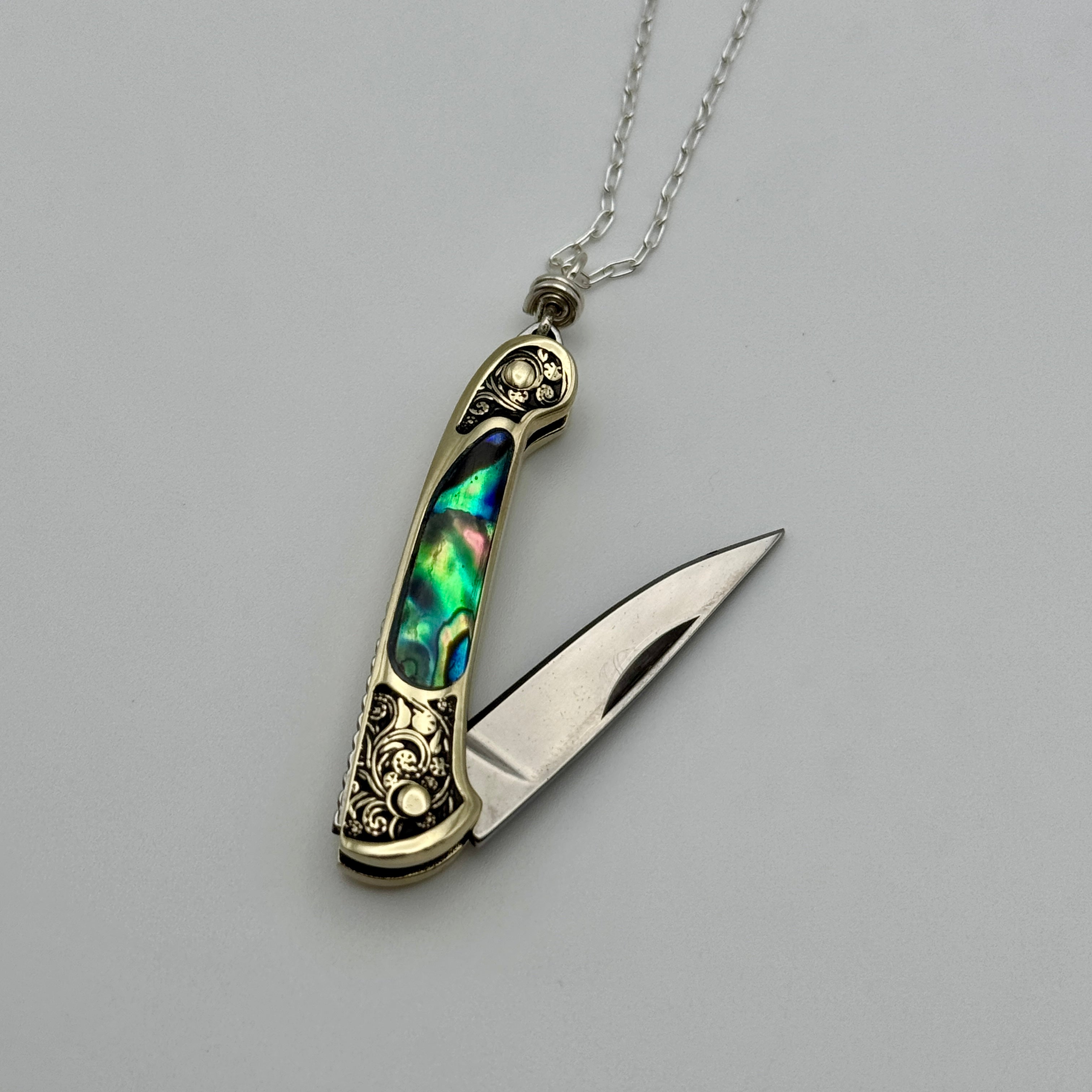 The Lennox Necklace by Fluff Hardware features a decorative pocket knife with a stainless steel blade, gold-tone floral-engraved handle, and iridescent inlay, styled as a utility pendant on a silver chain against a white background.
