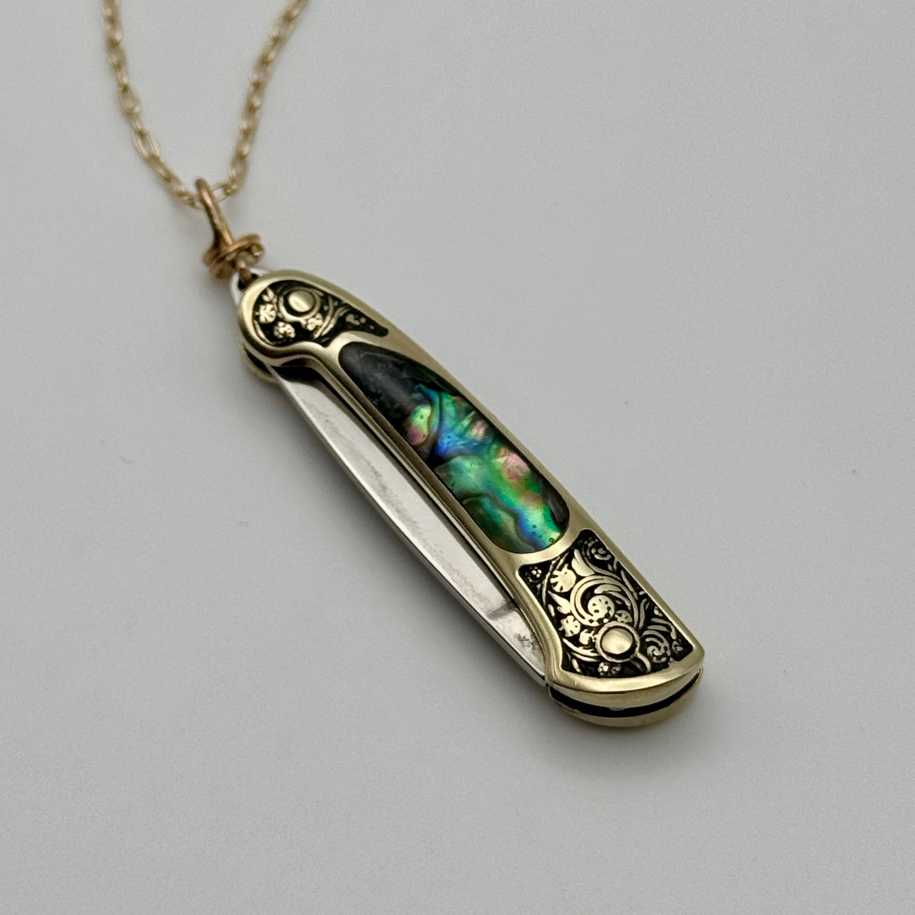 The Lennox Necklace by Fluff Hardware is a gold multi-tasking pendant shaped like a pocketknife with ornate engravings and an iridescent abalone inlay on the handle, shown against a plain light background.