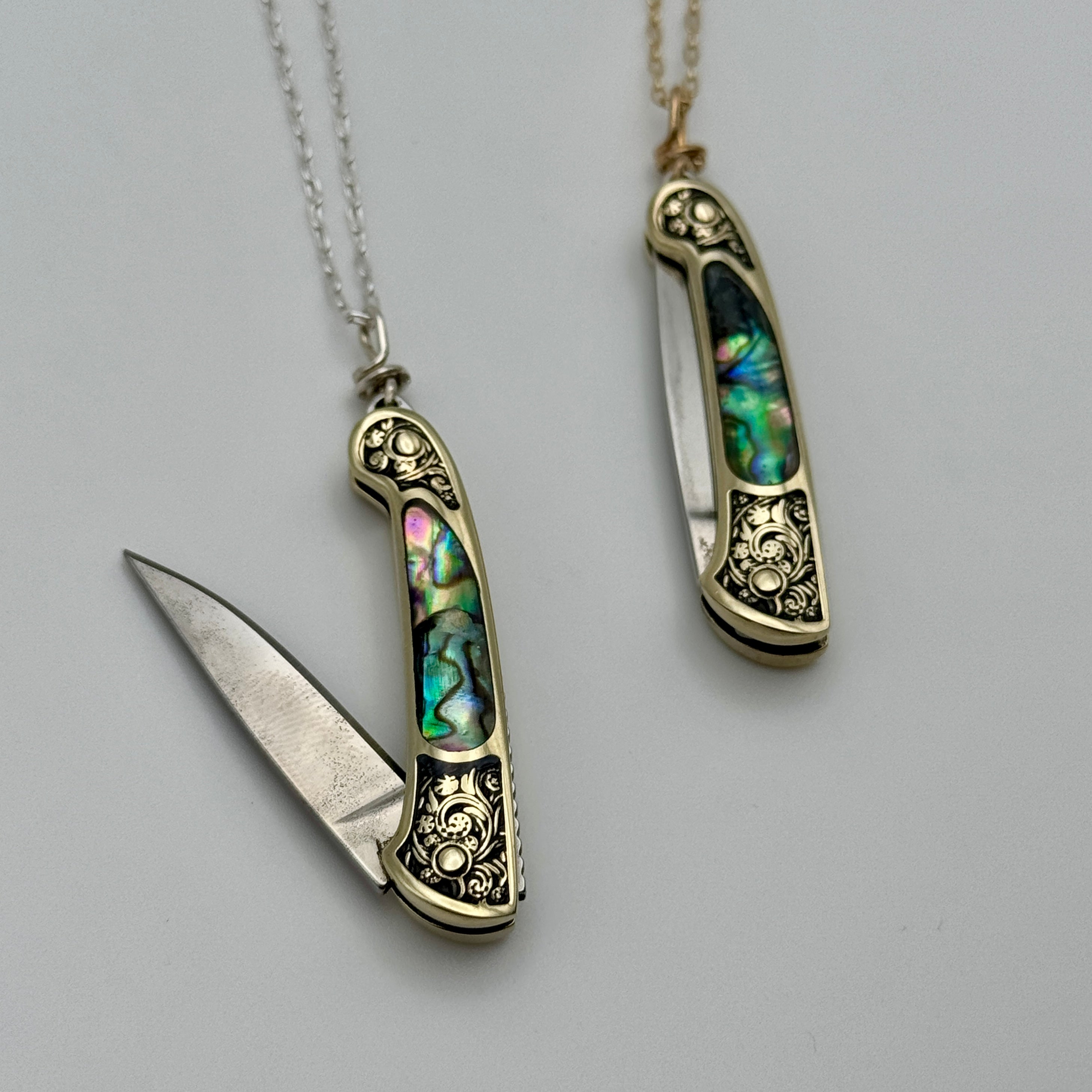 Two Lennox Necklaces by Fluff Hardware—ornate pocket knives with abalone shell inlays—hang on chains as unique functional jewelry; one knife is open, the other closed, each displayed on a plain background.