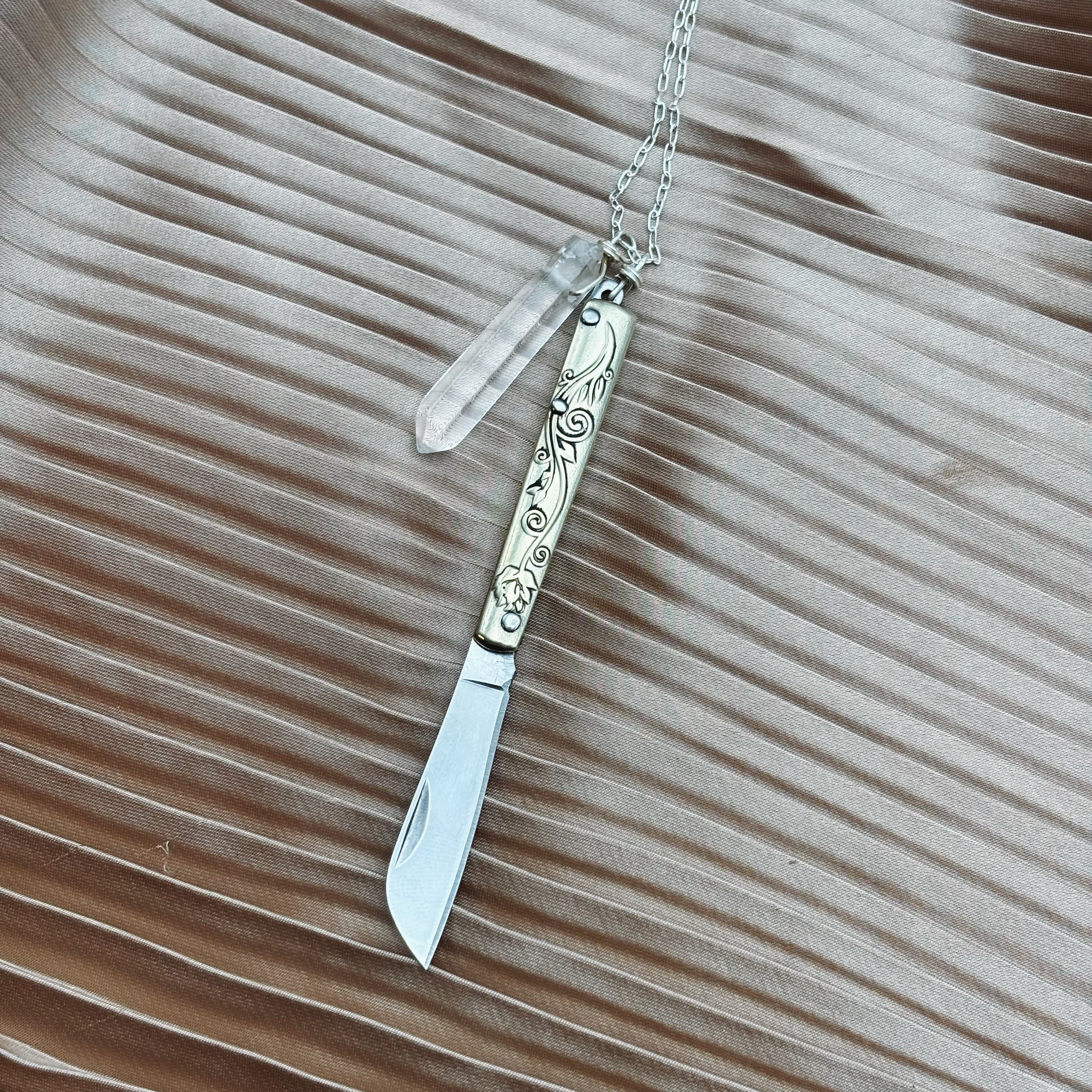 The Amelia Necklace by Fluff Hardware features a silver folding knife with an engraved handle and a quartz crystal pendant on a chain, shown against a textured beige surface.