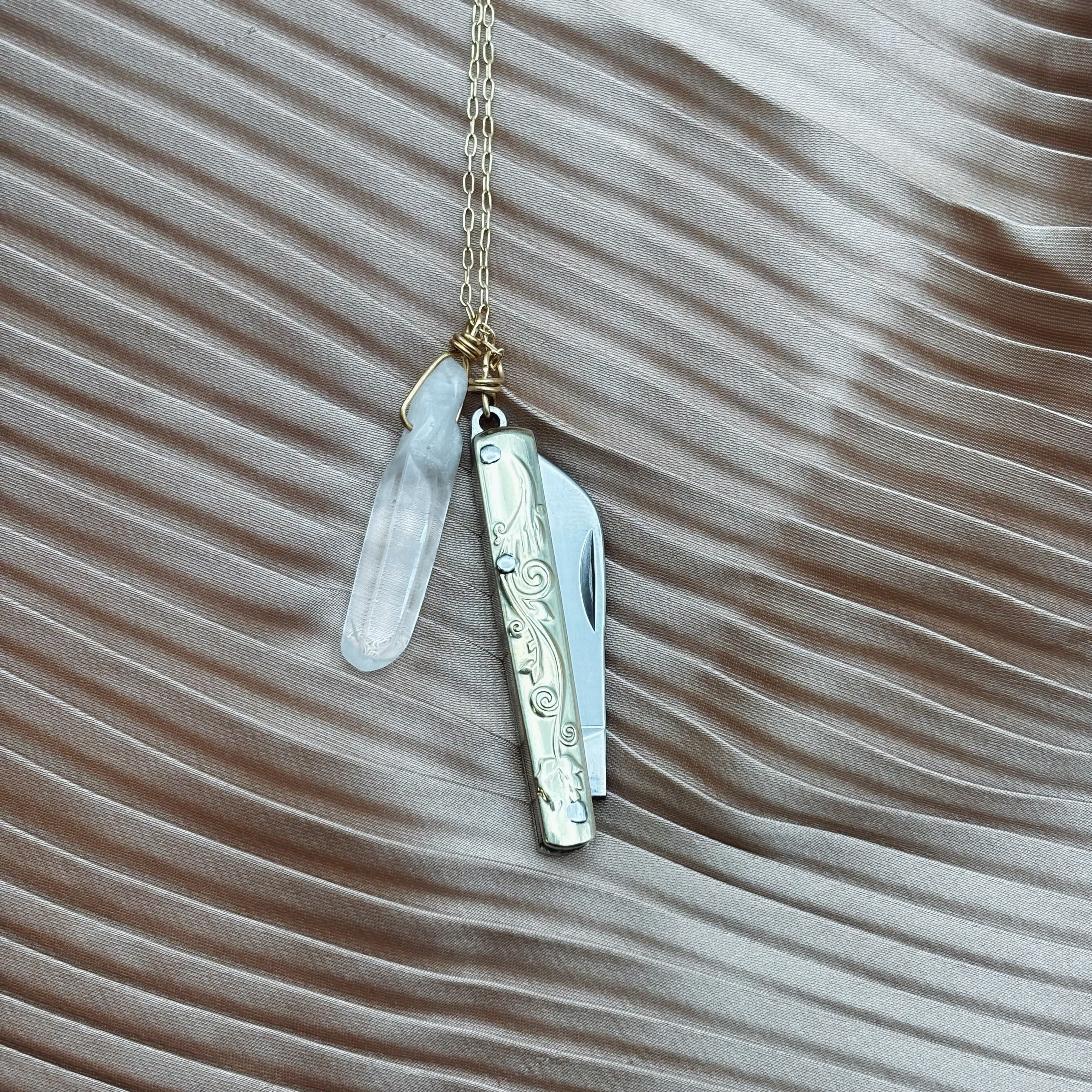 The Amelia Necklace by Fluff Hardware, featuring a gold chain with a quartz crystal and silver utility pendant, rests on silky light brown fabric with wave patterns.