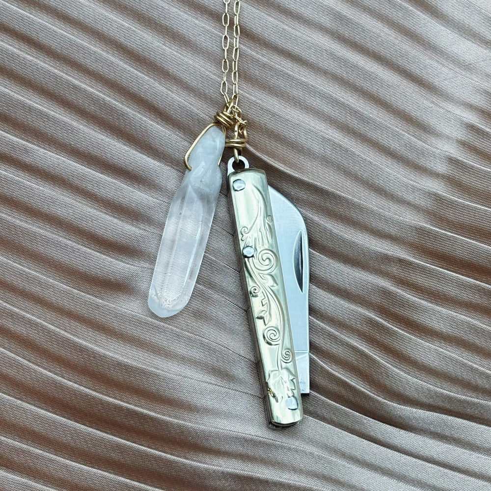 The Amelia Necklace by Fluff Hardware, featuring a gold chain with a quartz crystal and silver utility pendant, rests on silky light brown fabric with wave patterns.