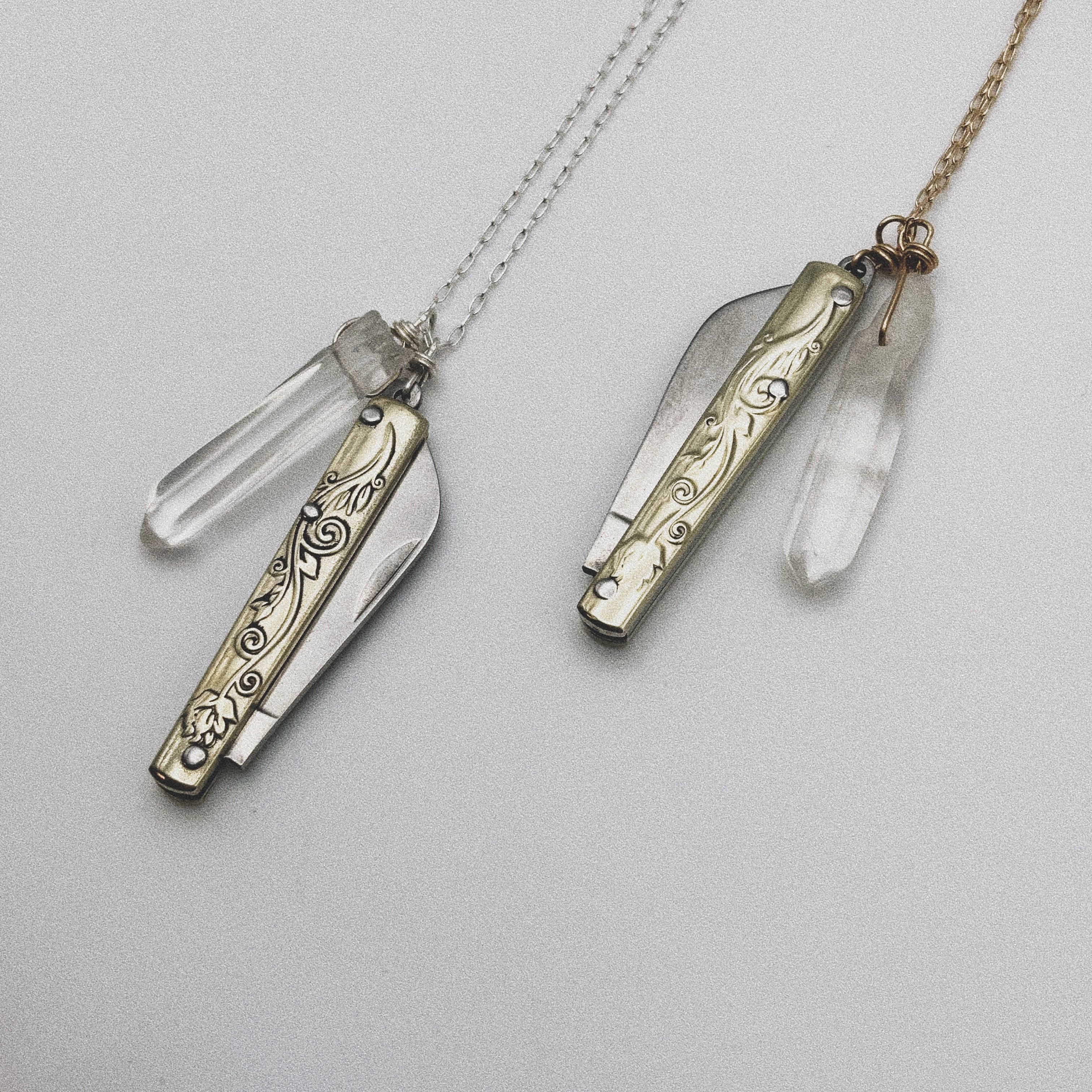 The Fluff Hardware Amelia Necklaces feature ornate, engraved utility pocketknife pendants with clear quartz crystal accents. Displayed on plain backgrounds, one has a silver chain, the other gold—unique, functional jewelry pieces.