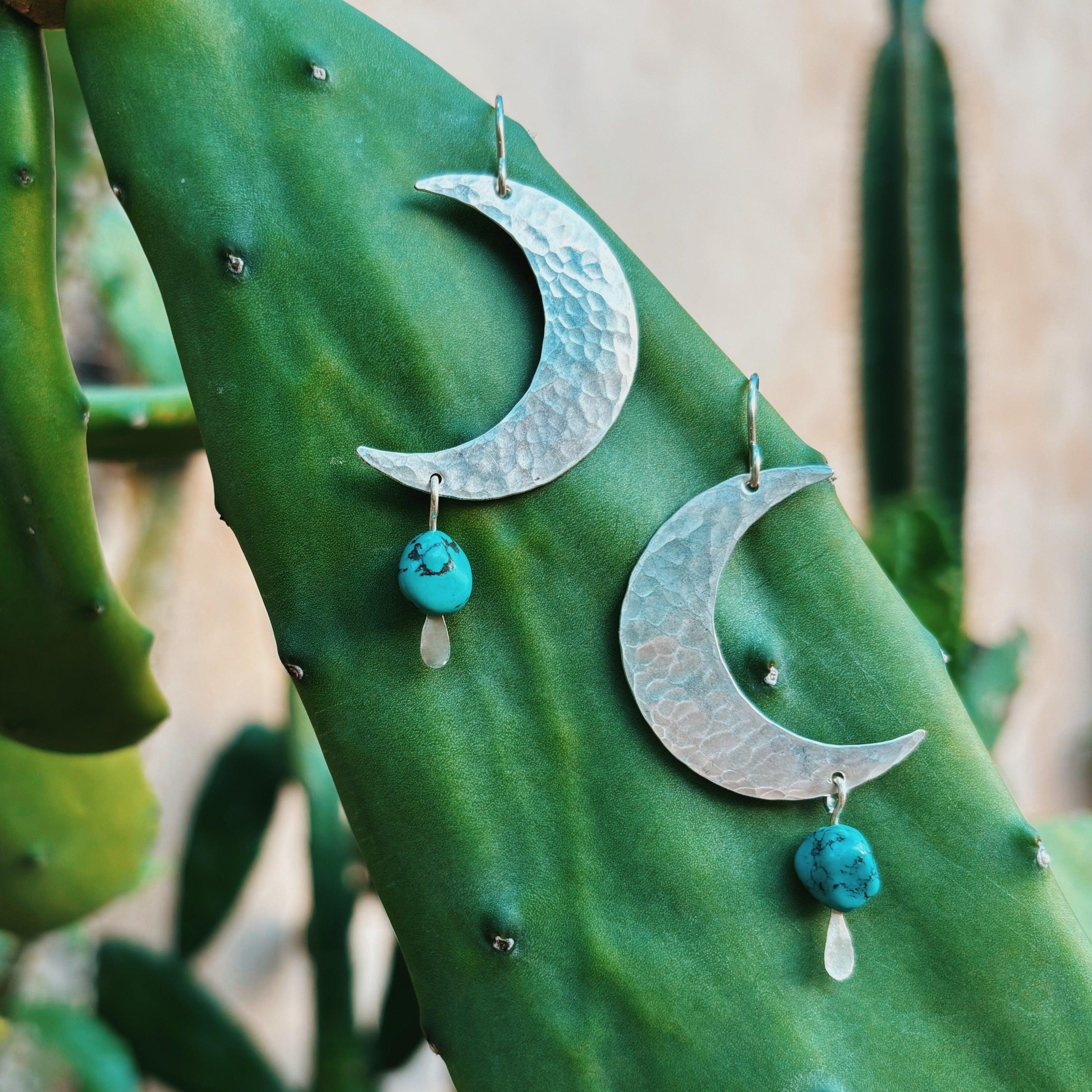 Fluff Hardwares Turquoise Moon Earrings feature hammered silver moons with dangling turquoise beads and white drops, displayed on a cactus leaf—perfect hypoallergenic jewelry for anyone who loves unique style.