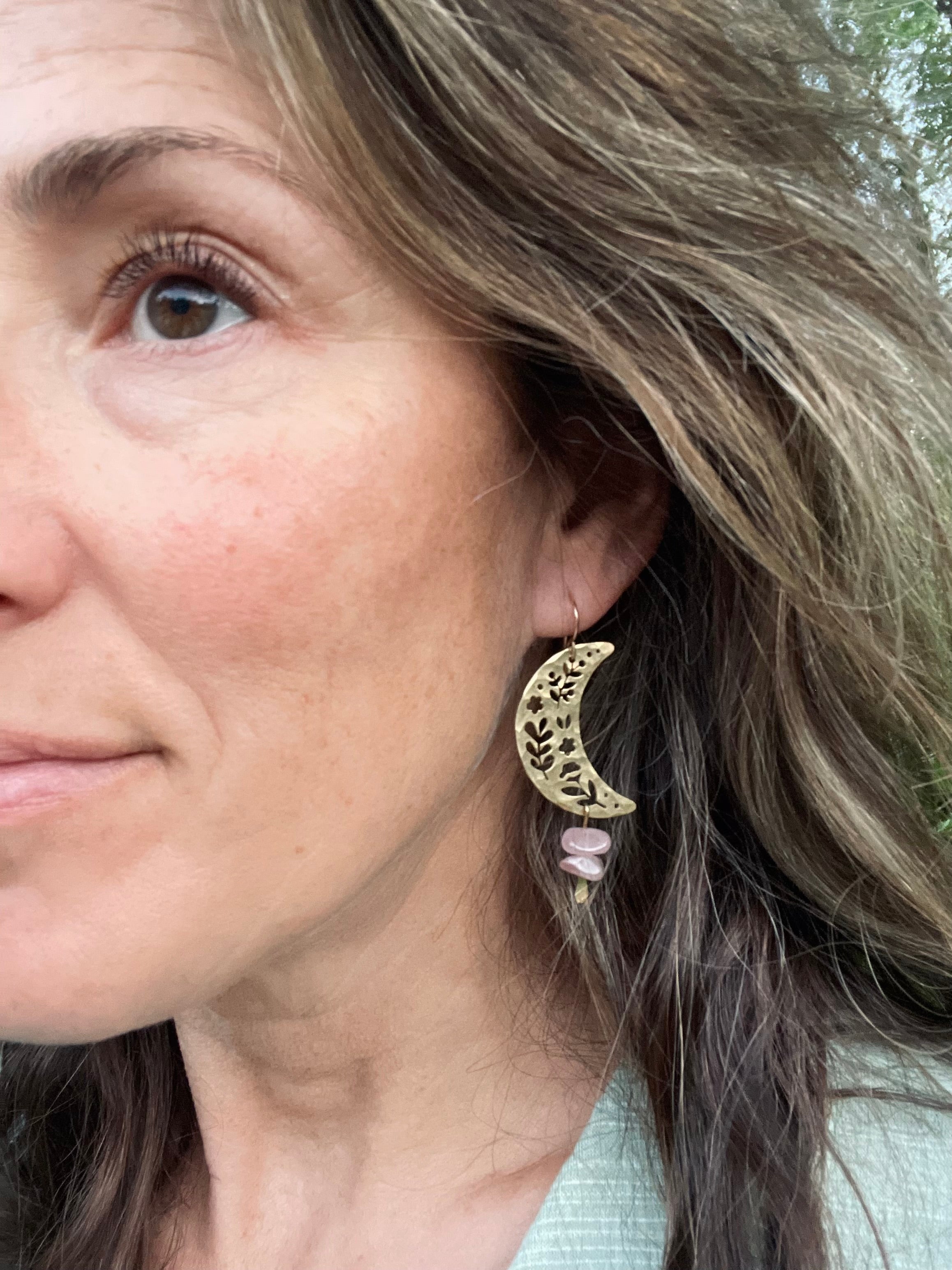 A woman with light skin, brown eyes, and wavy brown hair showcases Fluff Hardware’s Moon Garden Earrings—large, gold filled crescent moons with ornate cut-out details and rose quartz beads.