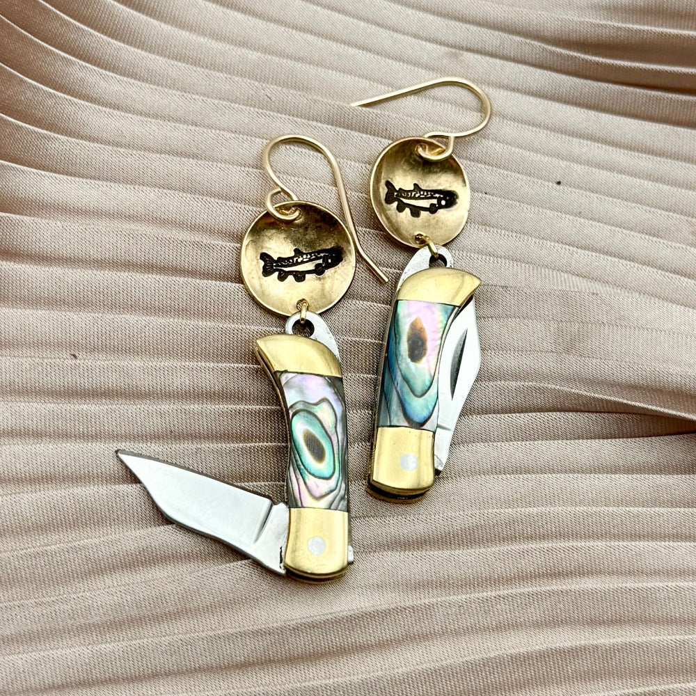 Fluff Hardwares Backcountry Earrings (Abalone) feature round gold discs stamped with fish and an abalone inlay pocket knife pendant earrings with gold accents, blending artistry and function atop beige pleated fabric.