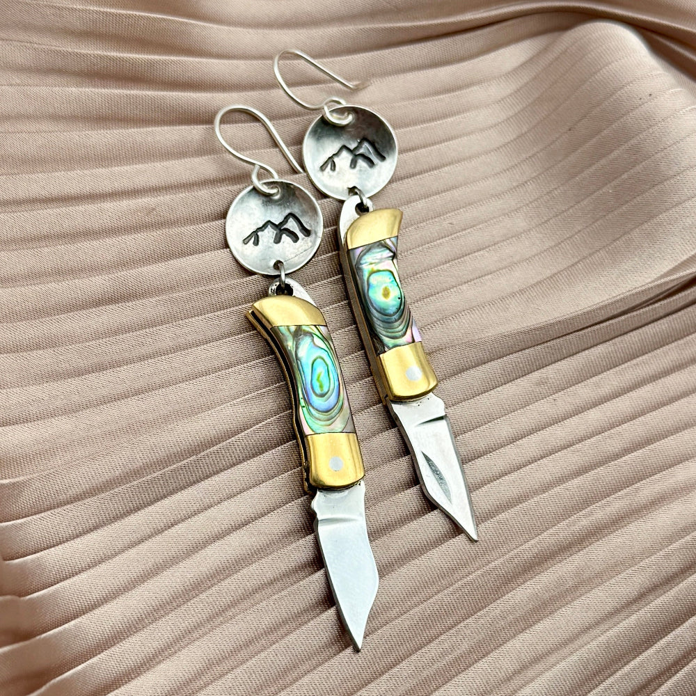 Fluff Hardwares Backcountry Earrings (Abalone) feature round silver discs stamped with mountain and an abalone inlay pocket knife pendant earrings with gold accents, blending artistry and function atop beige pleated fabric.