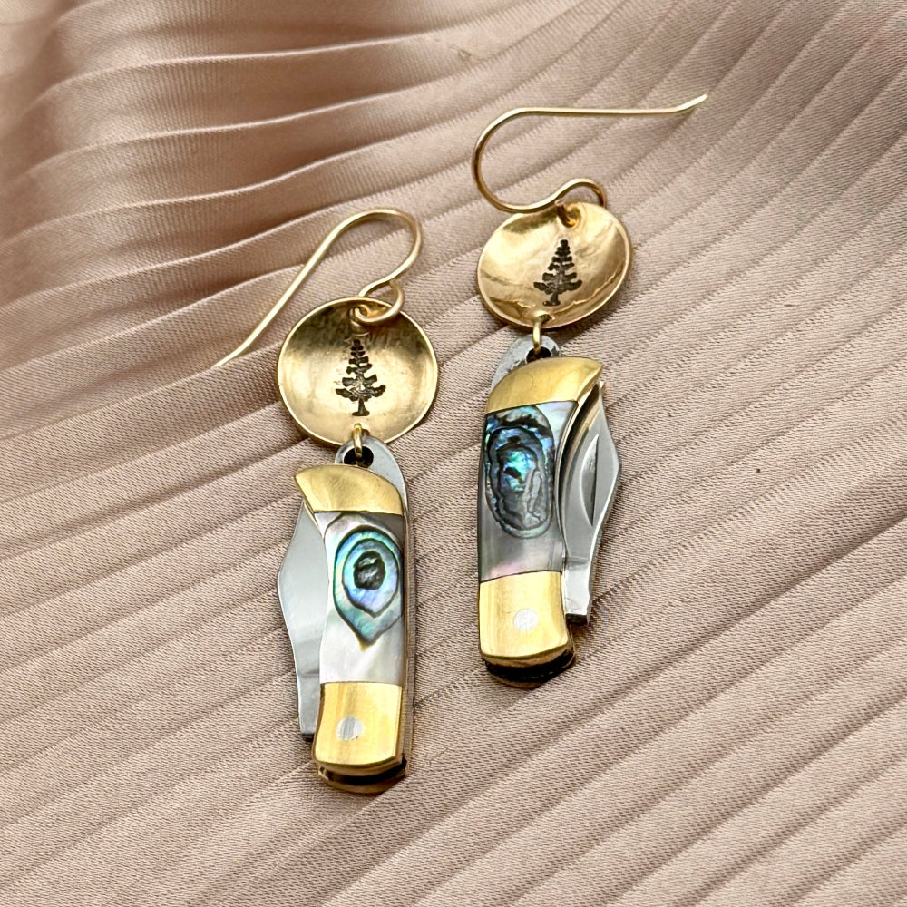 Fluff Hardwares Backcountry Earrings (Abalone) feature round gold discs stamped with tree and an abalone inlay pocket knife pendant earrings with gold accents, blending artistry and function atop beige pleated fabric.