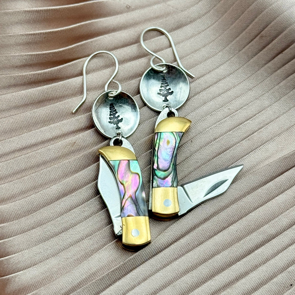 Fluff Hardwares Backcountry Earrings (Abalone) feature round silver discs stamped with tree and an abalone inlay pocket knife pendant earrings with gold accents, blending artistry and function atop beige pleated fabric.