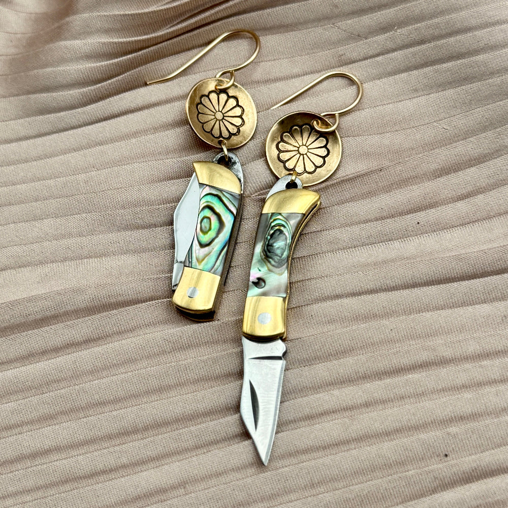 Fluff Hardwares Backcountry Earrings (Abalone) feature round gold discs stamped with flower and an abalone inlay pocket knife pendant earrings with gold accents, blending artistry and function atop beige pleated fabric.