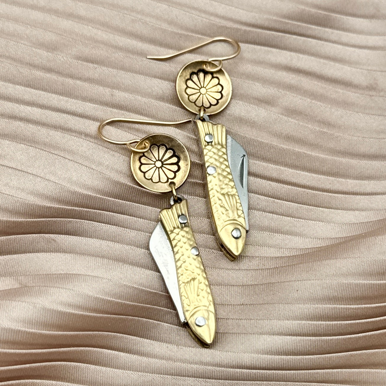 The Backcountry Feeesh Earrings by Fluff Hardware are gold and sterling silver pocketknife earrings with floral engravings, blending charm and function—shown here on pleated beige fabric.
