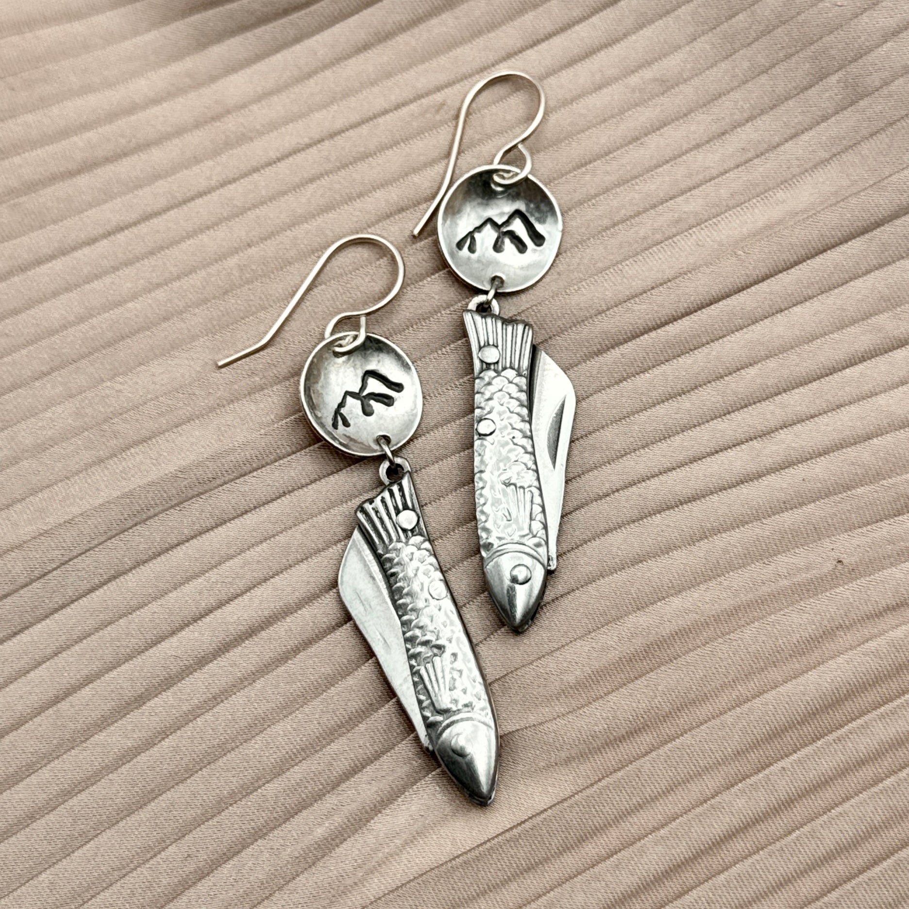 The Backcountry Feeesh Earrings by Fluff Hardware feature silver fish-shaped pendants with circular, engraved mountain tops, blending artistry and everyday elegance. Displayed on textured beige fabric.