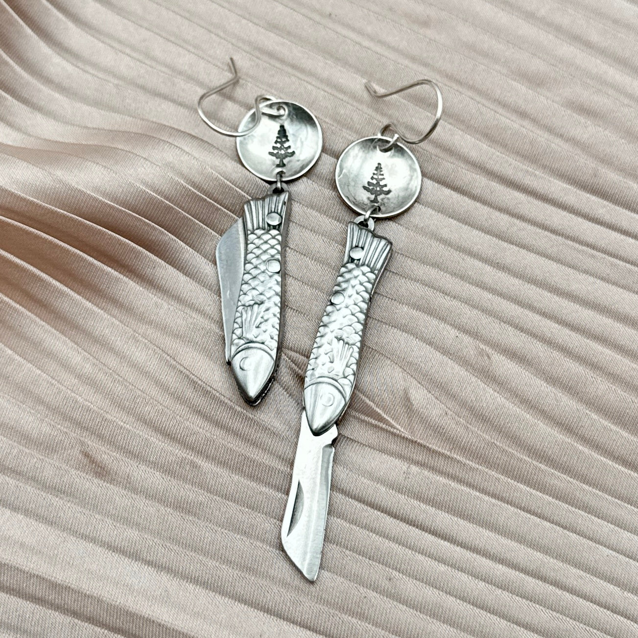 The Backcountry Feeesh Earrings by Fluff Hardware are silver or gold earrings with miniature fish pocketknife charms, each hanging from a round gold disc engraved with a pine tree, displayed on textured beige fabric.