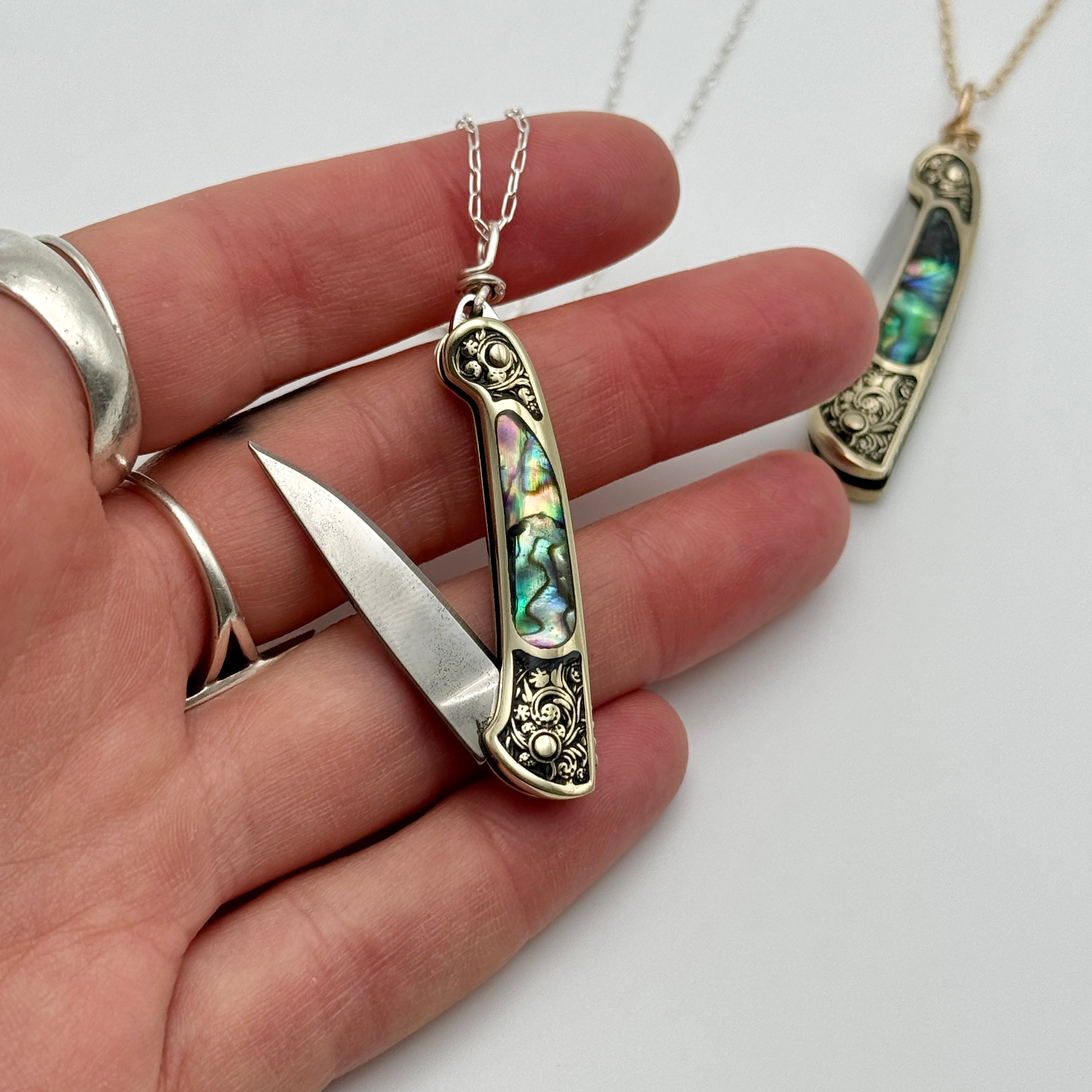 A hand holds the Fluff Hardware Lennox Necklace, a pocketknife utility pendant with abalone inlay and ornate metalwork on a silver chain. Another Lennox Necklace on a gold chain is softly blurred in the background.