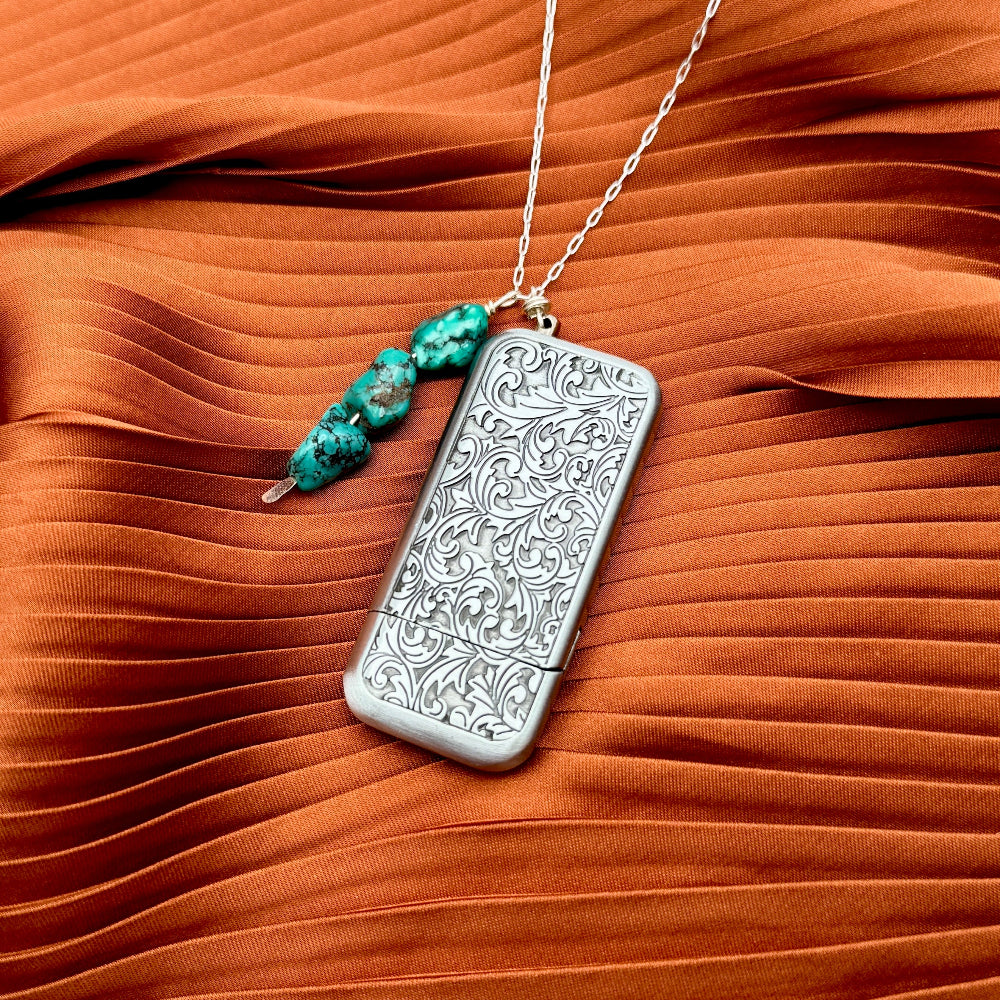 The Flicker Lighter Necklace by Fluff Hardware features a silver rectangular lighter with floral engravings and turquoise beaded