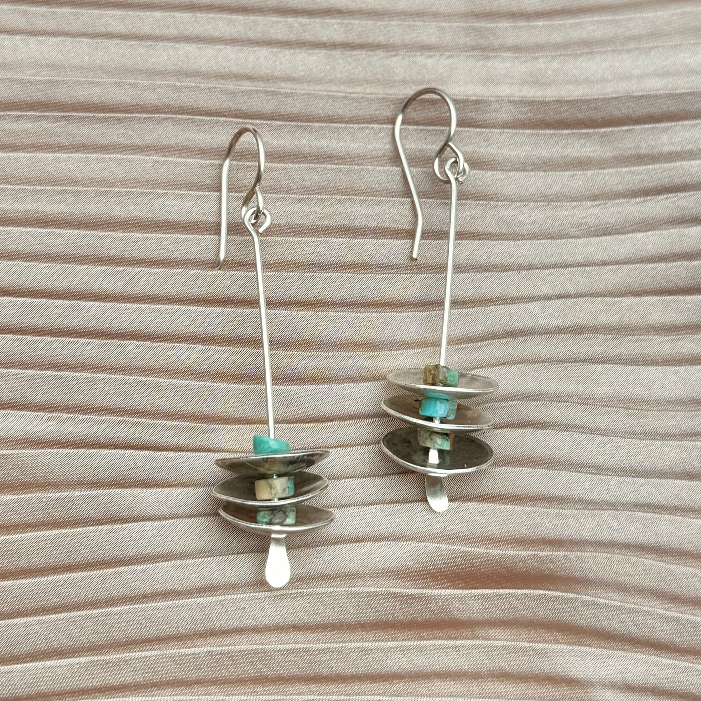 The Remains Earrings by Fluff Hardware feature three stacked sterling silver discs and small turquoise beads on long hypoallergenic hooks, displayed on a wavy beige fabric background. Perfect for sensitive ears.