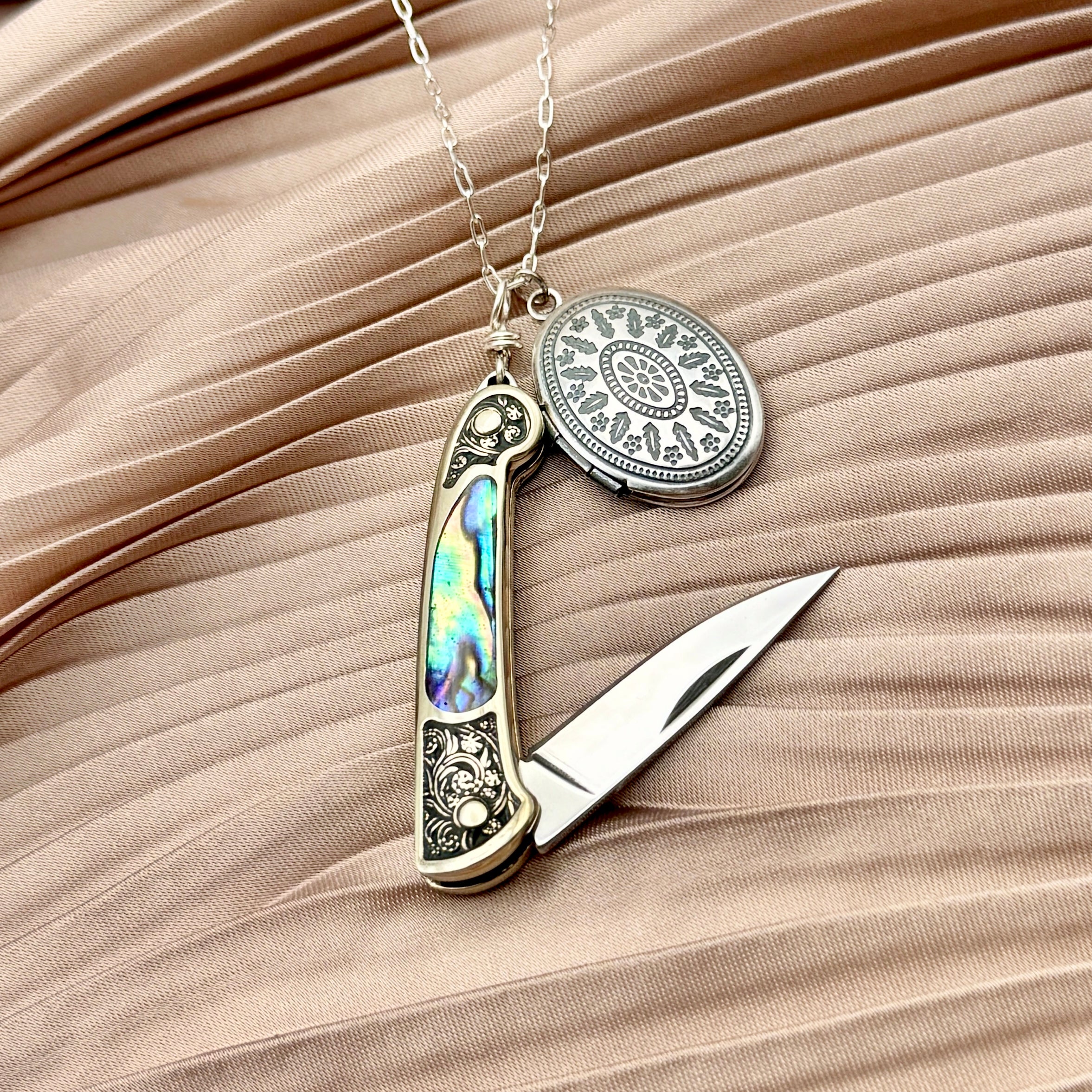 The Fluff Hardware Keepsake Necklace features a silver oval locket and a small abalone-inlaid pocket knife, creating an elegant piece of functional jewelry displayed on pleated beige fabric.