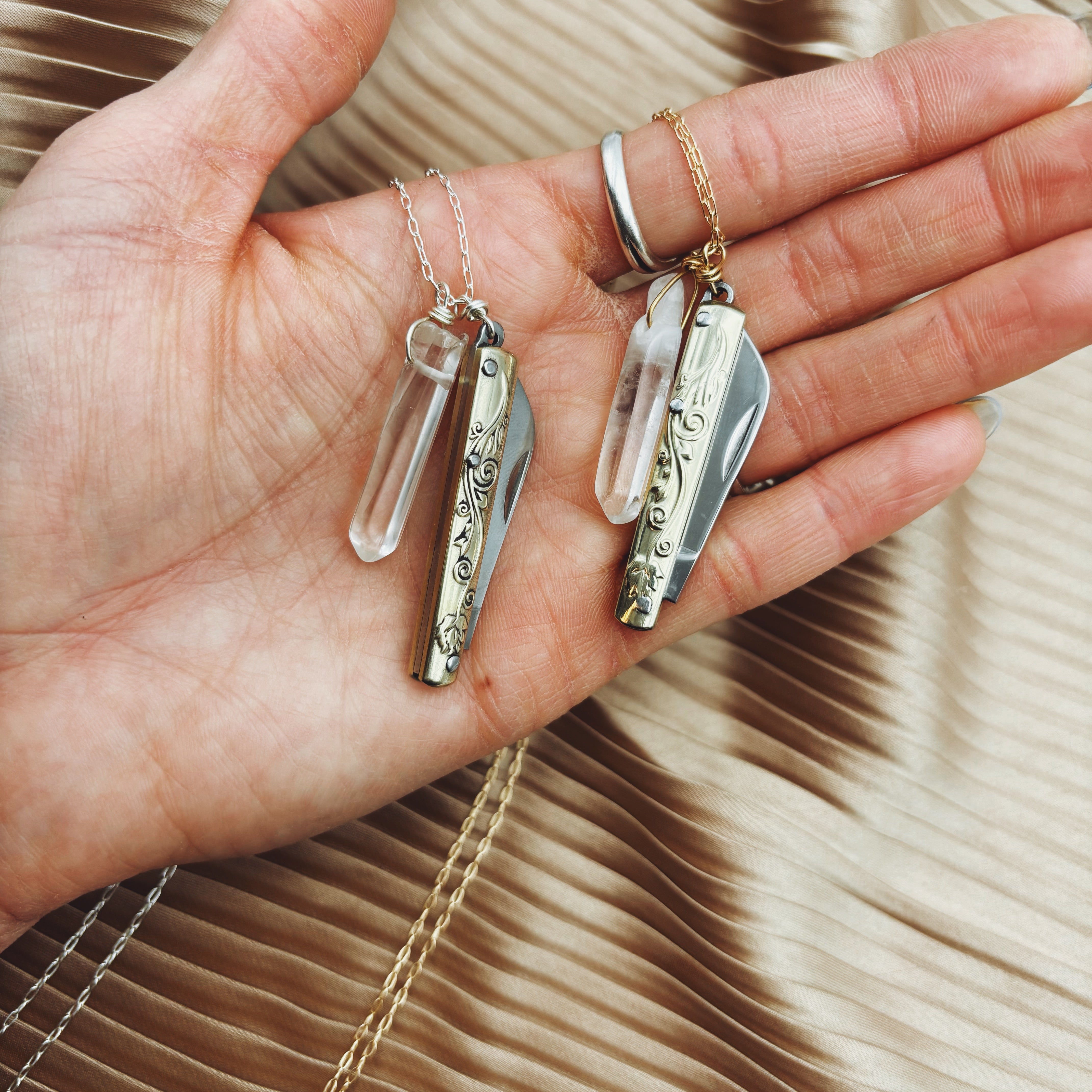 A hand holds two Fluff Hardware Amelia Necklaces, each with an ornate silver pocket knife and a quartz crystal pendant, set against pleated beige fabric.