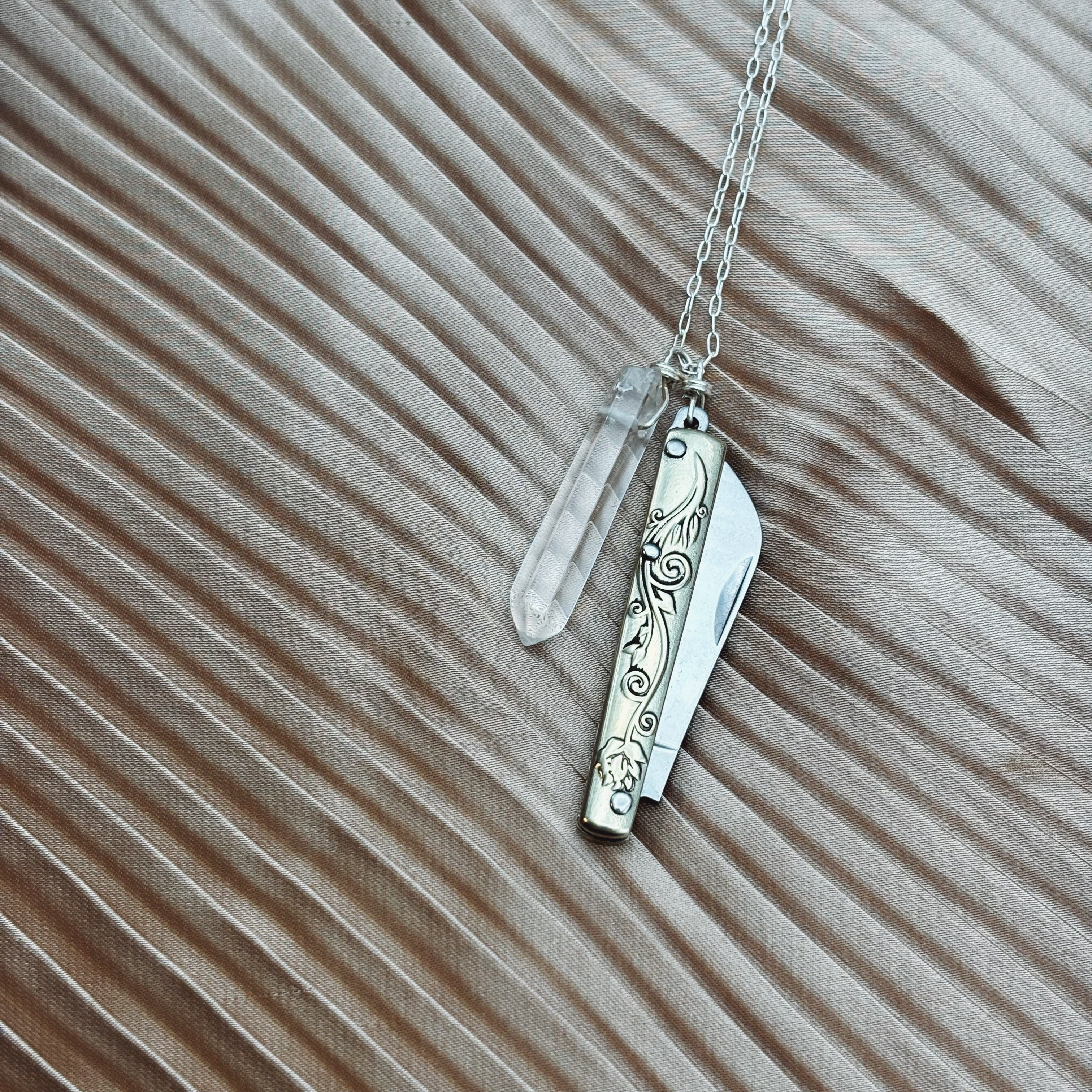 The Amelia Necklace by Fluff Hardware is a silver piece featuring a pocket knife charm and clear quartz crystal pendant, displayed on pleated beige fabric.