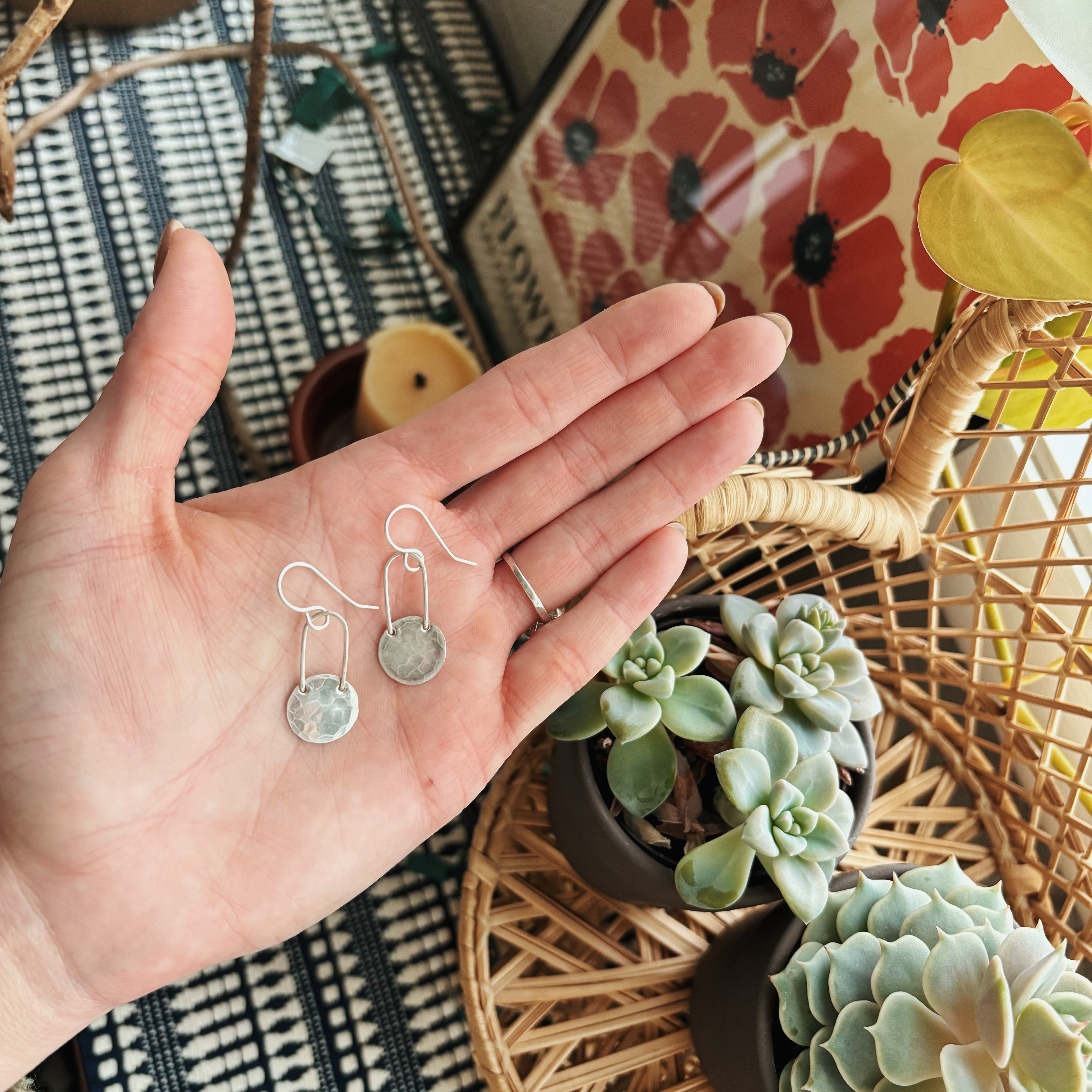 A hand holds Fluff Hardwares Fallen Moon Earrings—silver, hypoallergenic dangle jewelry—above a patterned surface, with succulents, a candle, and floral artwork in the background.