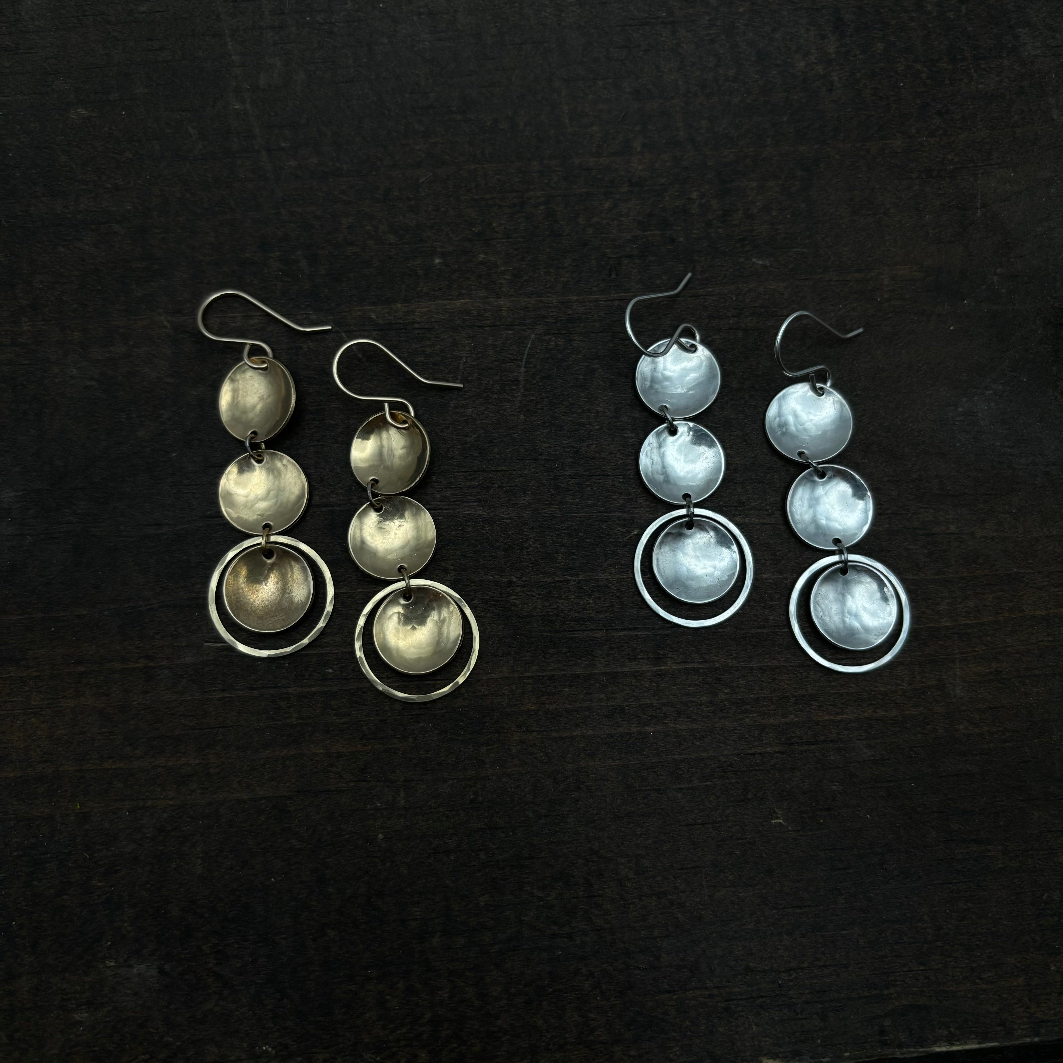 Two pairs of Fluff Hardware Rubble Earrings on a dark surface: one set in gold and one in silver, both with segmented circle pendants and rings, crafted from hypoallergenic materials.