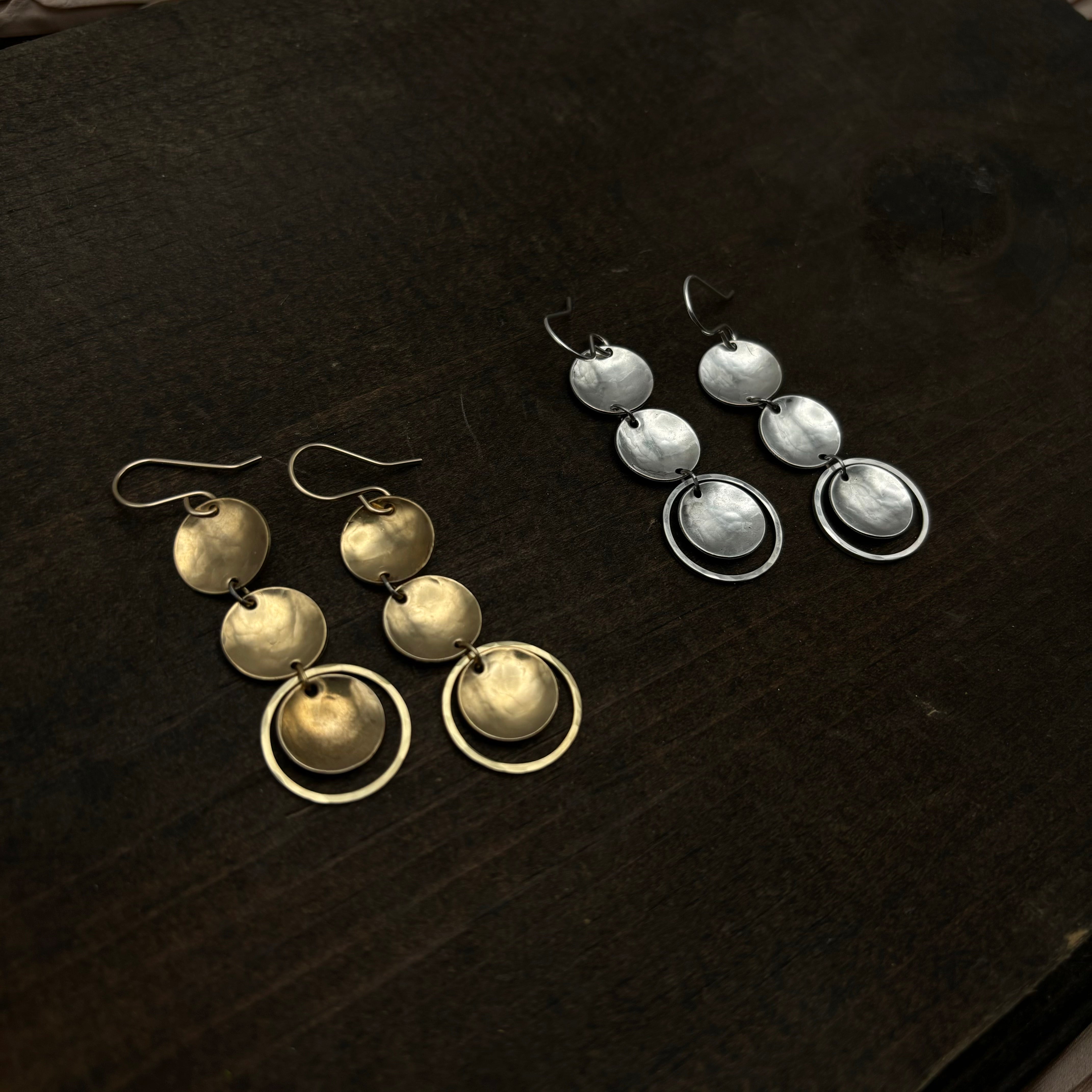 Two pairs of Rubble Earrings by Fluff Hardware rest side by side on a dark surface: one in gold with three discs and a ring, and the other with sterling silver ear wires. Both are hypoallergenic dangle earrings.
