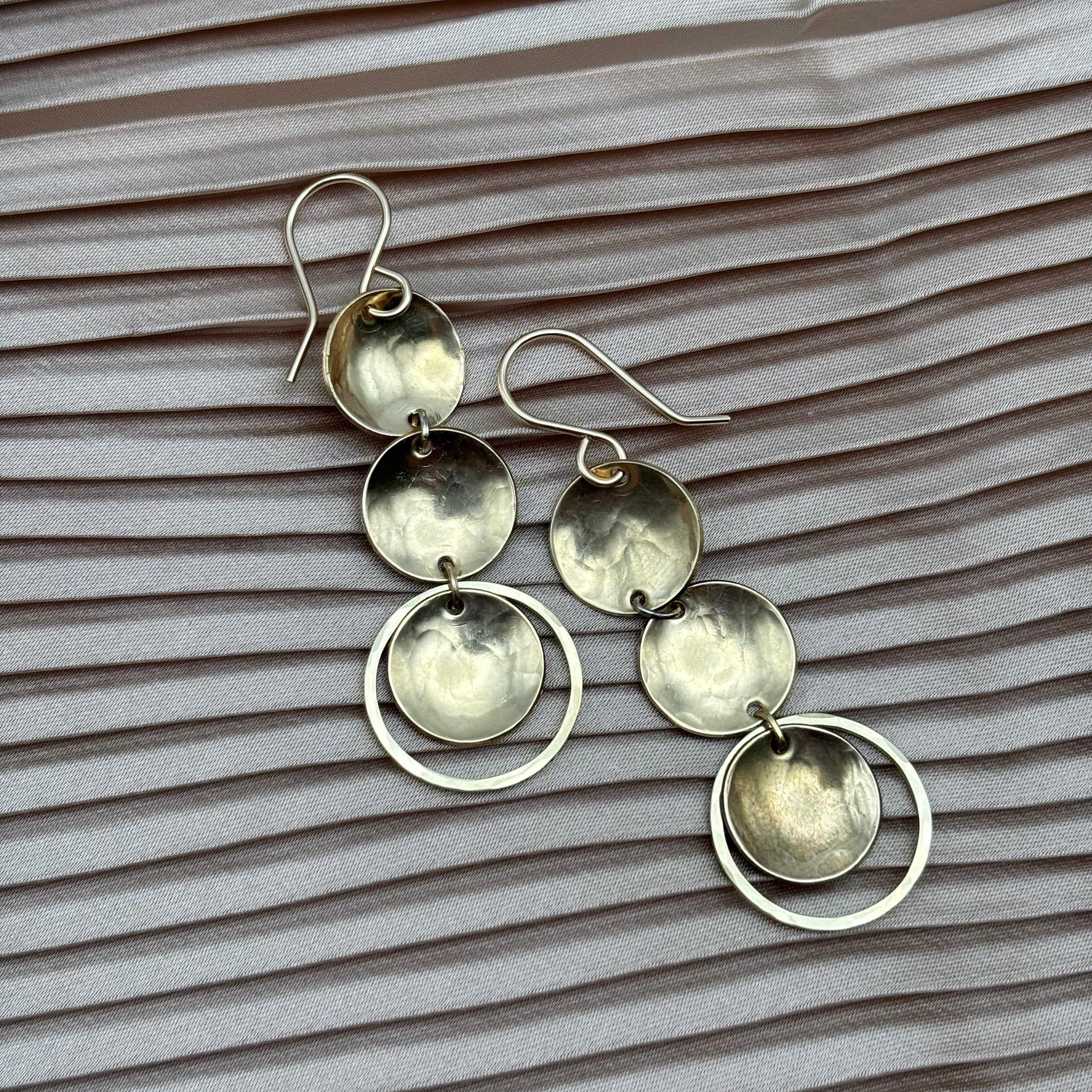 The Fluff Hardware Rubble Earrings are silver dangle earrings with three linked circular discs and an open ring, finished with sterling silver ear wires, shown against textured light pink fabric.