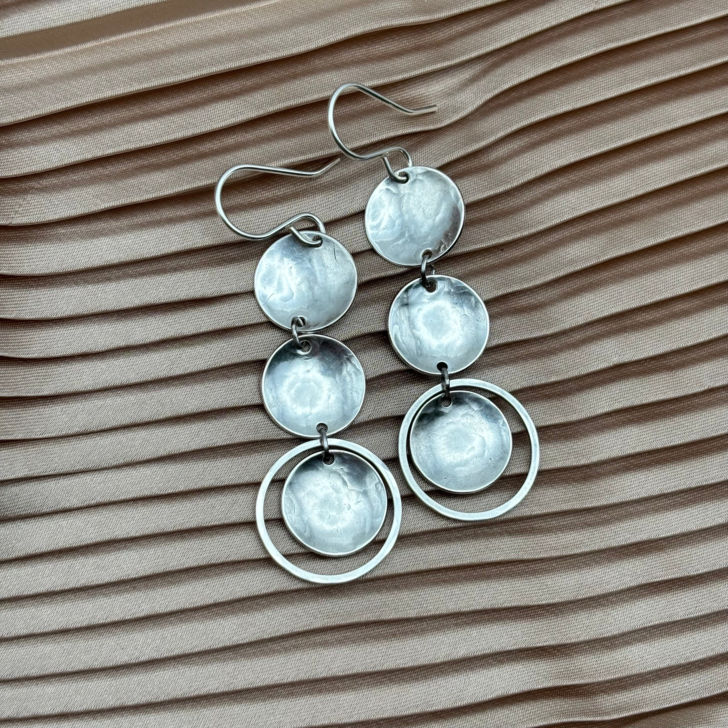 The Rubble Earrings by Fluff Hardware feature three hammered discs and two overlapping hoops in silver, hanging from sterling silver ear wires on pleated light brown fabric.