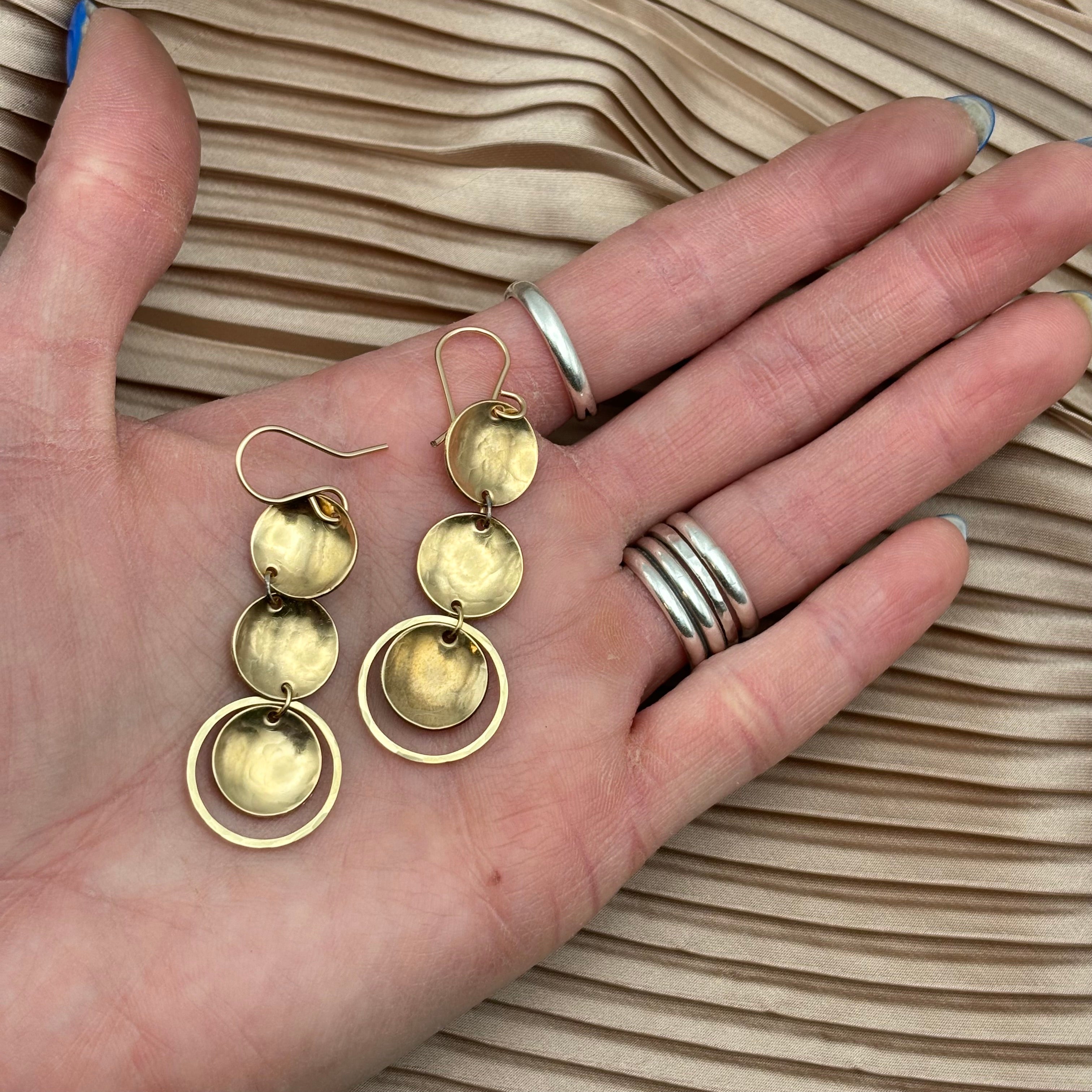 A hand displays Fluff Hardwares Rubble Earrings—gold dangle earrings with three hammered discs, two open circles, and sterling silver ear wires—paired with three silver rings against pleated beige fabric.