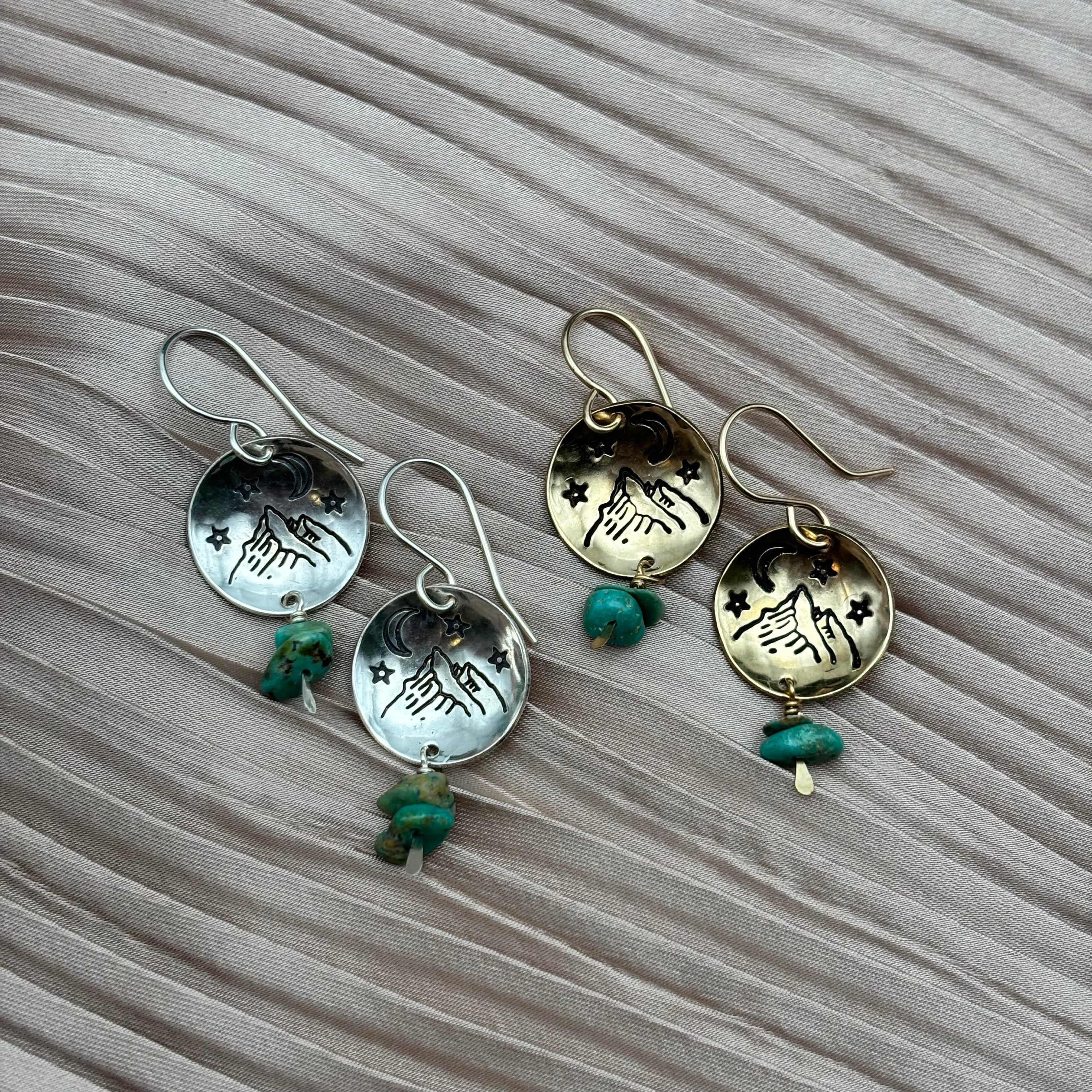 Four Starry Night Earrings by Fluff Hardware rest on pleated fabric. Each features a round hand-and-stars charm, hypoallergenic ear wires, and a green stone bead. Set includes two silver-toned and two gold-toned pairs.