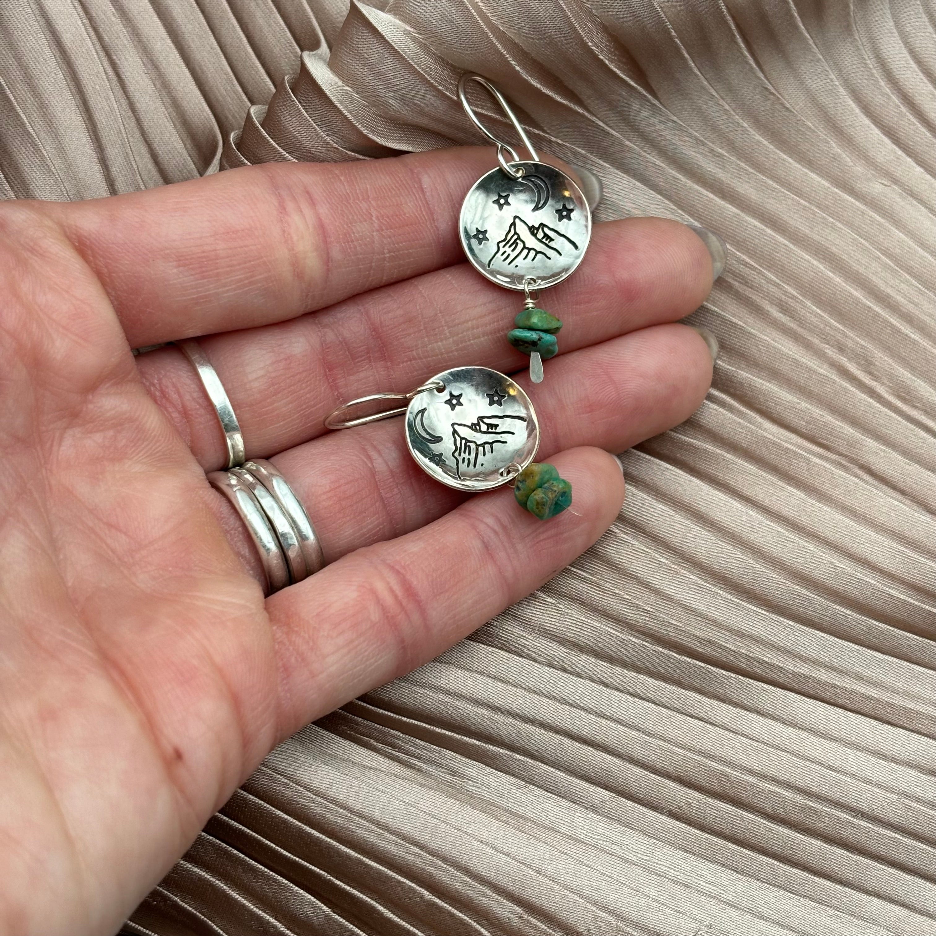 A hand holds Fluff Hardwares Starry Night Earrings—round silver pieces engraved with howling wolves, stars, and a crescent moon, each accented by a green bead and hypoallergenic wires—set against pleated beige fabric.