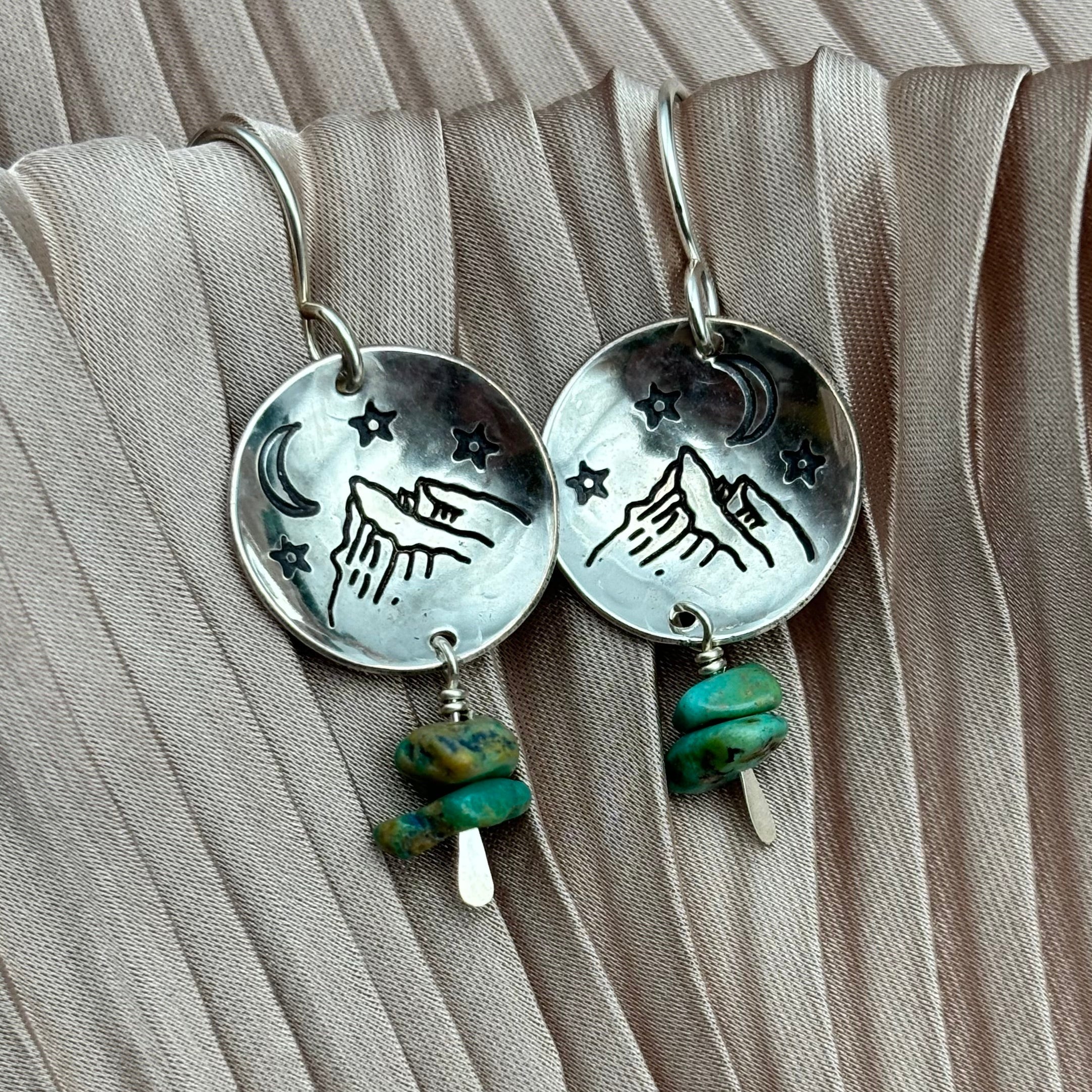 The Starry Night Earrings by Fluff Hardware feature round silver mountain-stamped pendants with engraved stars and a crescent moon, accented by two dangling turquoise beads. Hypoallergenic ear wires provide comfort. Shown on pleated beige fabric.