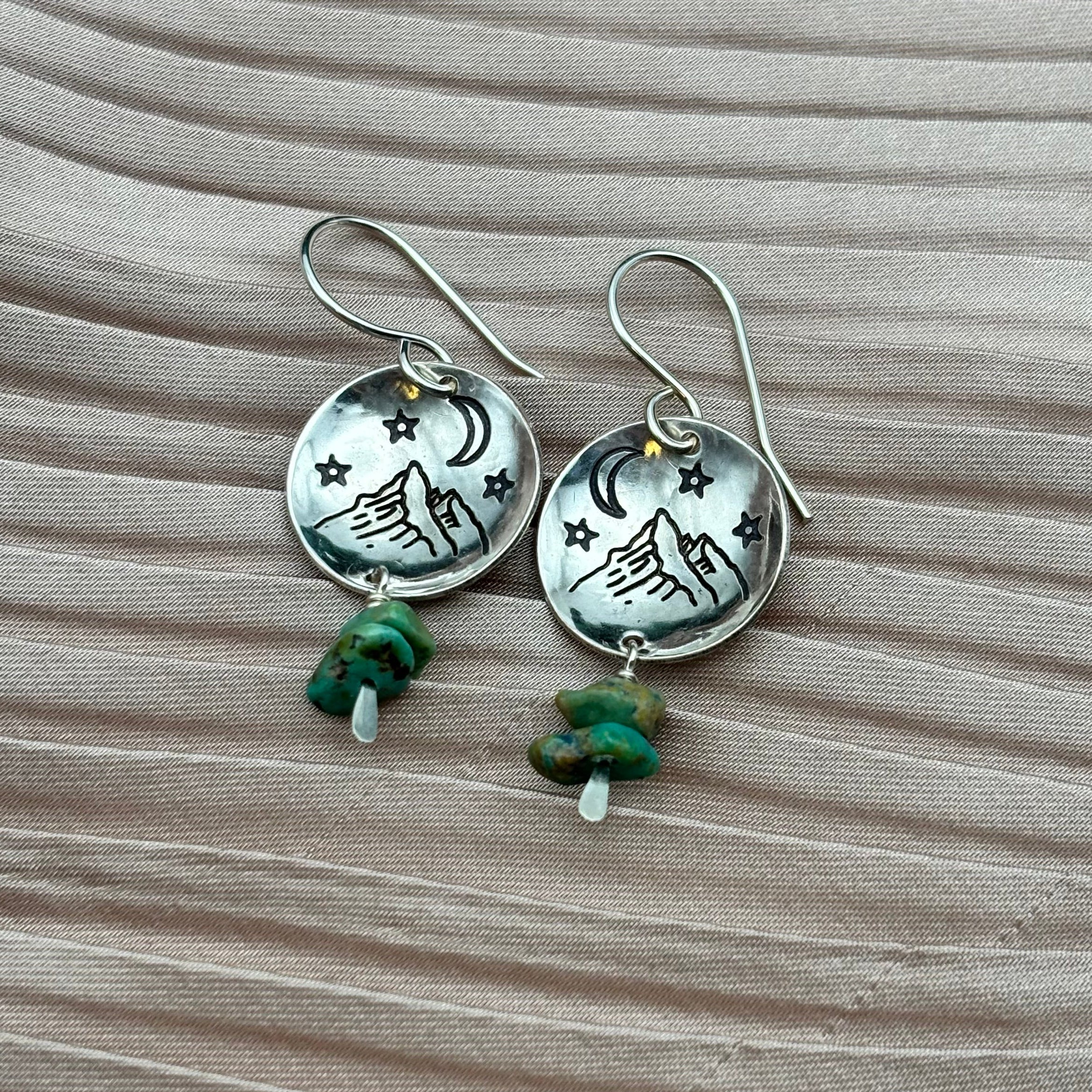 The Starry Night Earrings by Fluff Hardware feature silver mountain-stamped pendants with star and moon engravings, accented by green gemstones and mother of pearl beads, hanging from hypoallergenic wires on a textured backdrop.