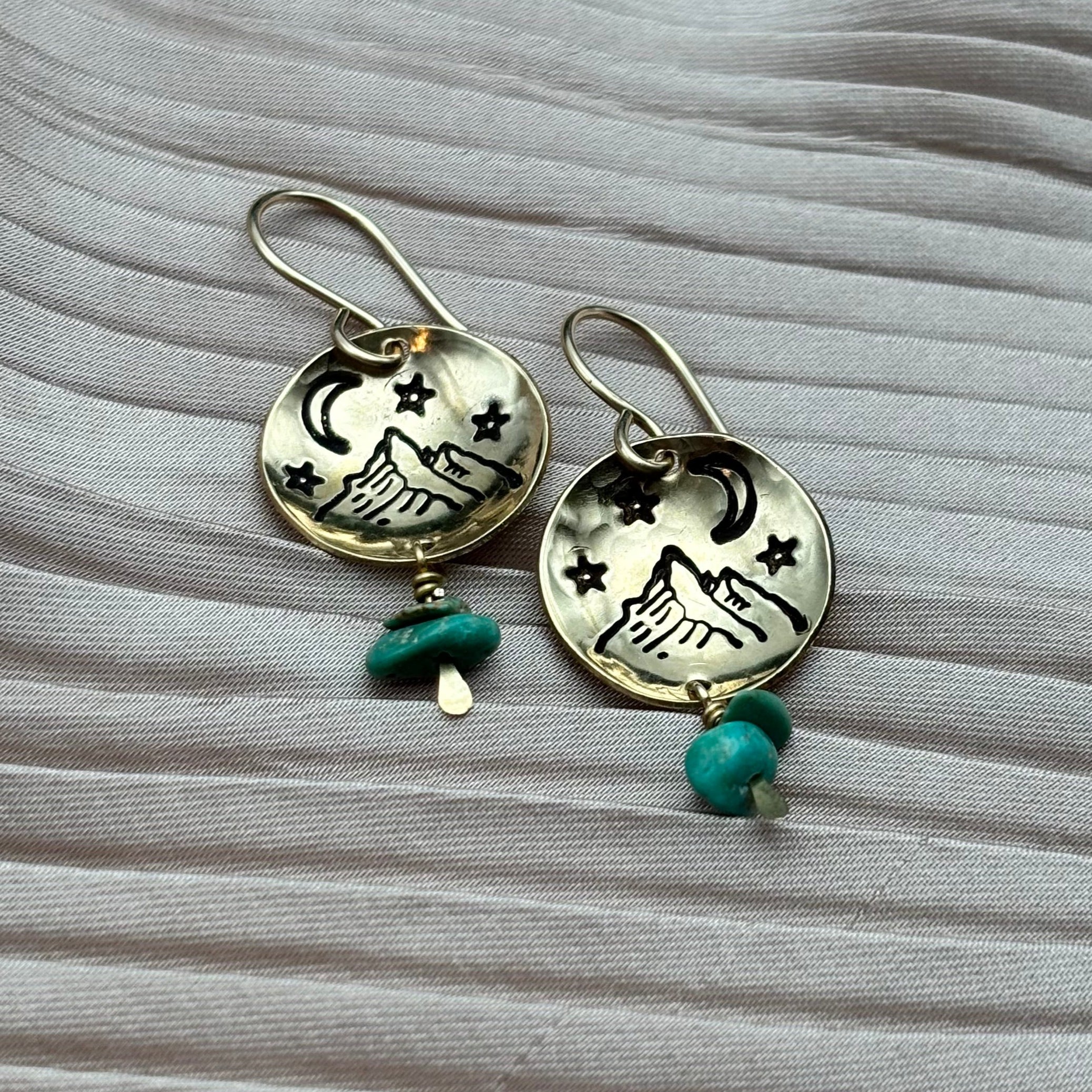 The Starry Night Earrings by Fluff Hardware feature round, etched howling wolves, stars, and crescent moons with turquoise, white beads, and mother of pearl accents, displayed on textured light grey fabric.