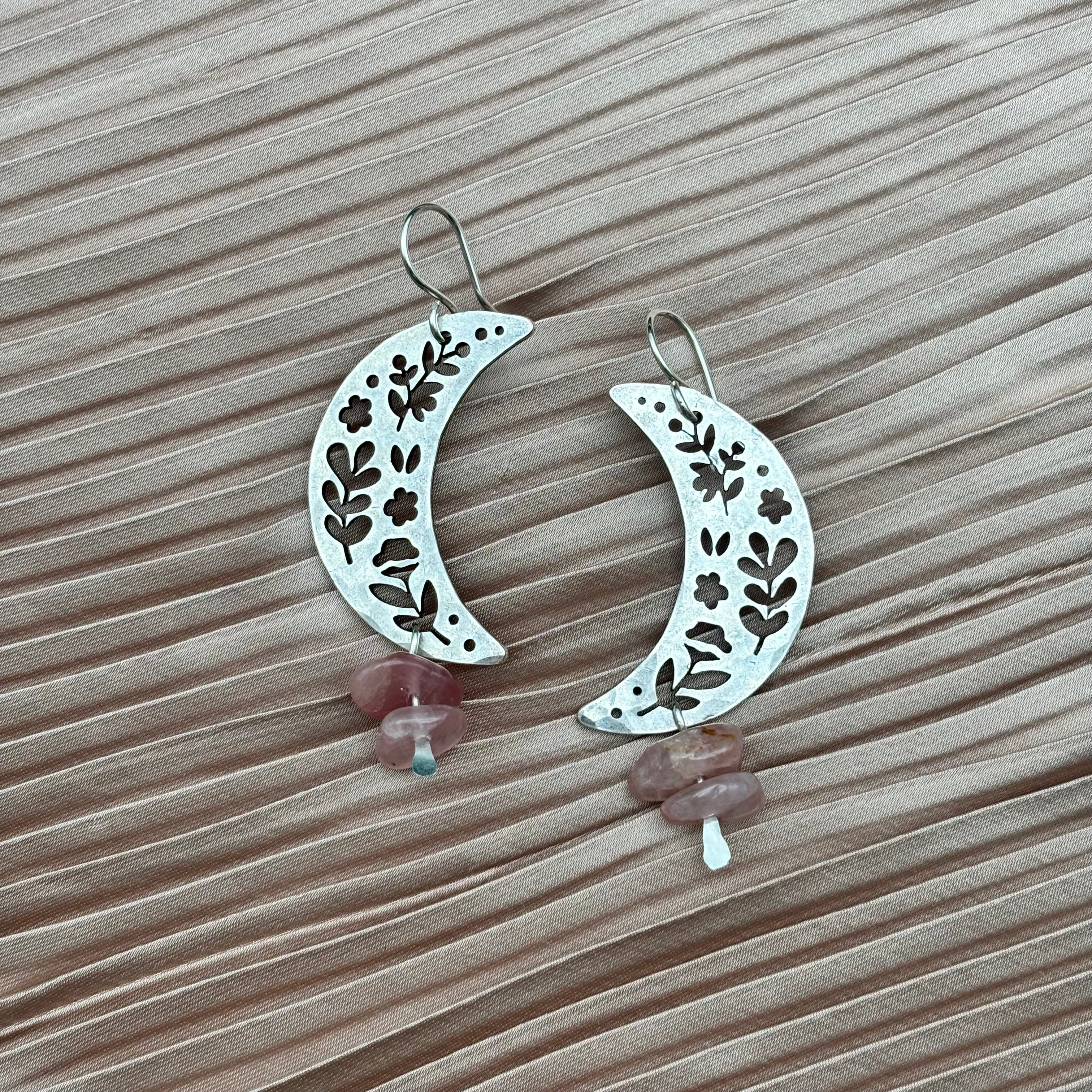 The Moon Garden Earrings by Fluff Hardware feature crescent moon shapes with floral cutouts and rose quartz beads, shown on a textured light pink fabric background.