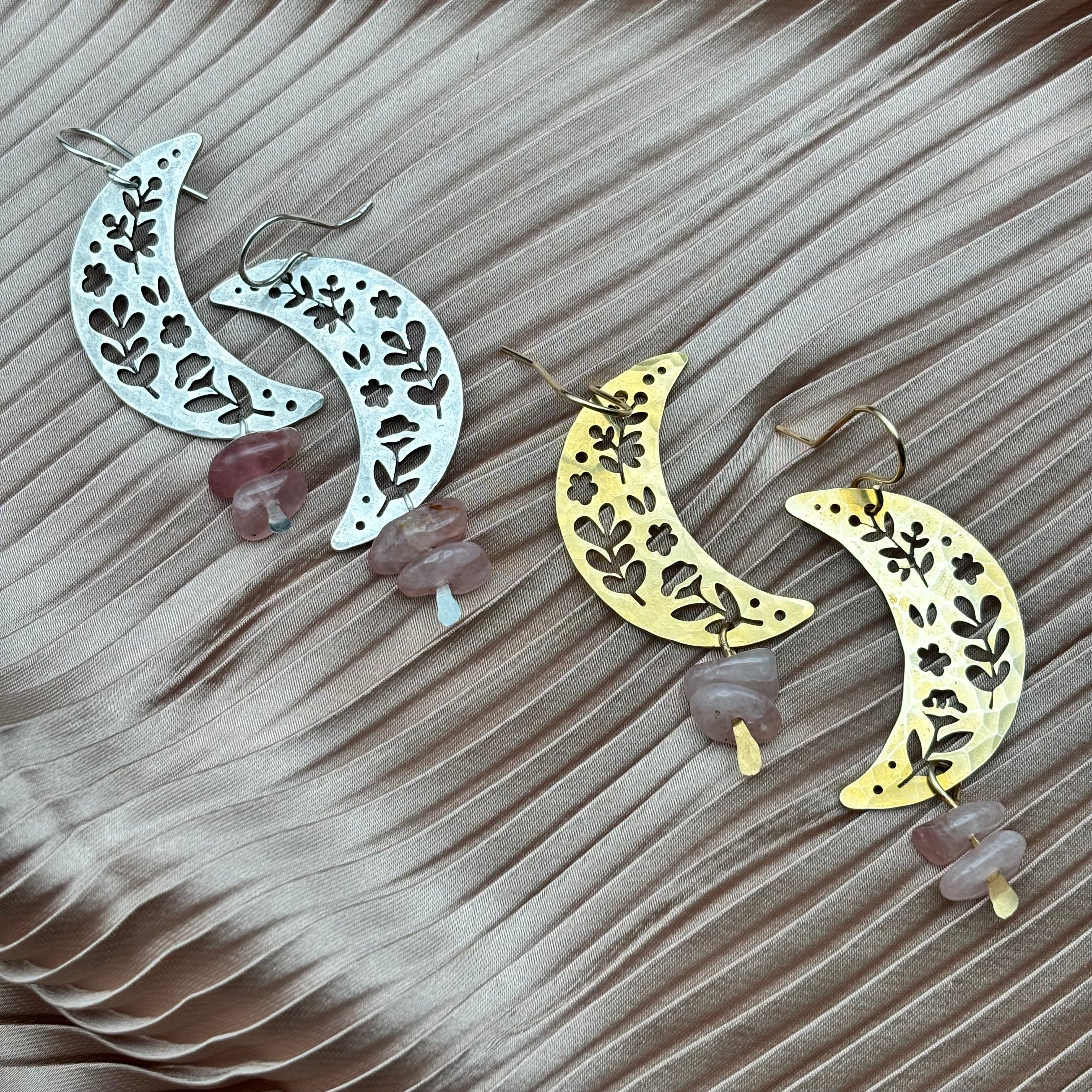 The Moon Garden Earrings by Fluff Hardware feature two crescent moon-shaped pairs—one silver, one gold—with floral cutouts and stacked rose quartz beads, displayed on pleated light pink fabric. A delicate, elegant style inspired by the moon.