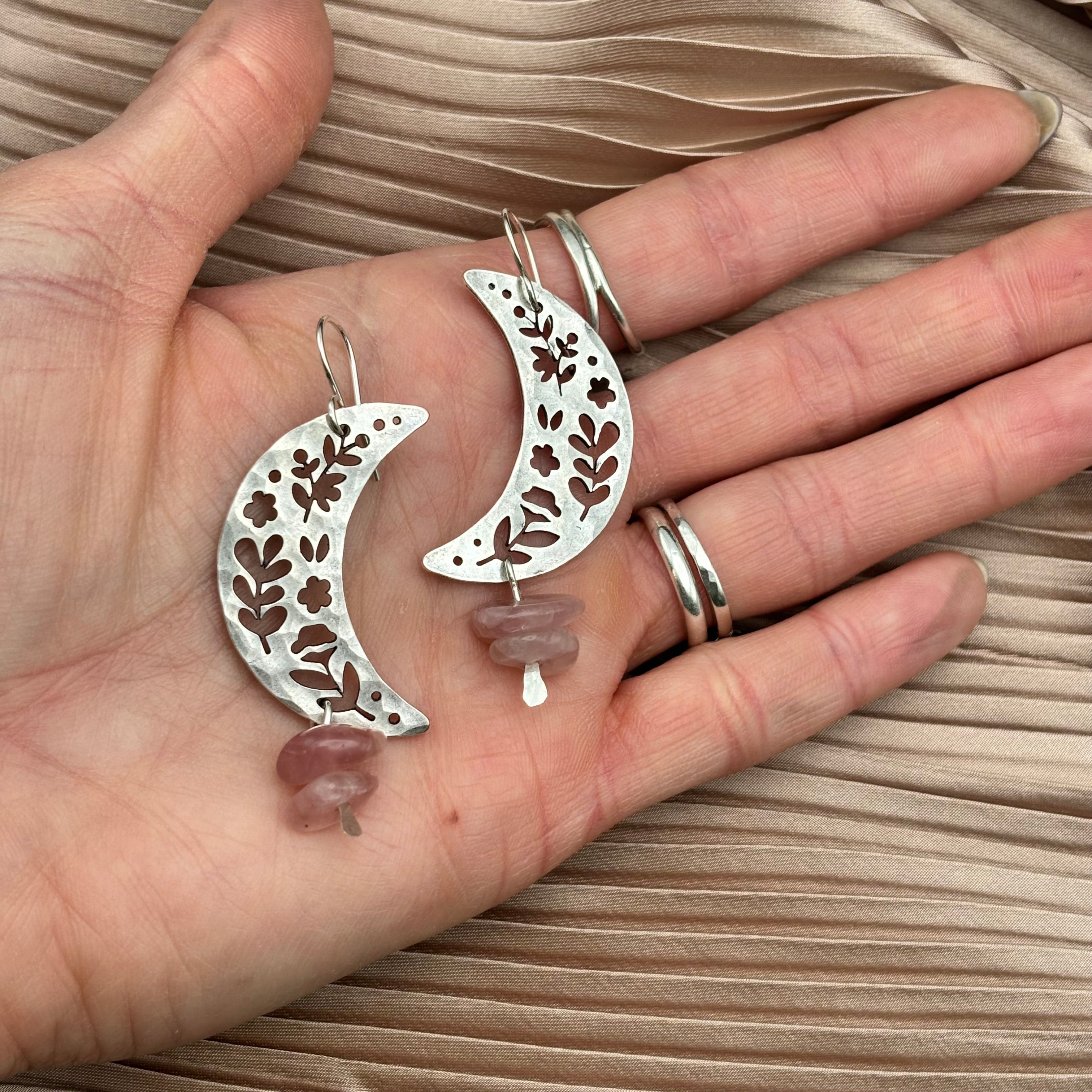 A hand holds Fluff Hardwares Moon Garden Earrings—crescent-shaped with floral cutouts and pink rose quartz beads—set against pleated beige fabric.