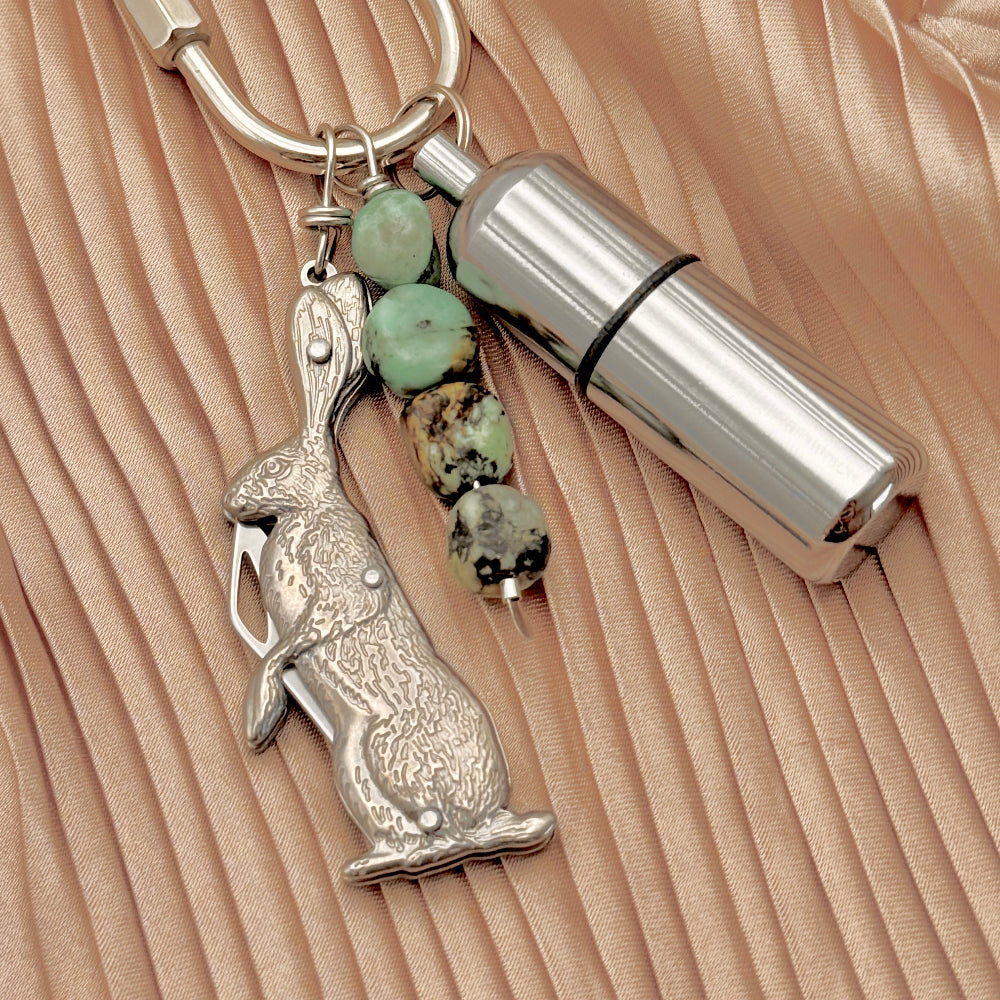A silver-toned metal keychain featuring a detailed rabbit charm, a cylindrical bottle lighter, and three four turquoise beads, displayed on a textured beige fabric background.