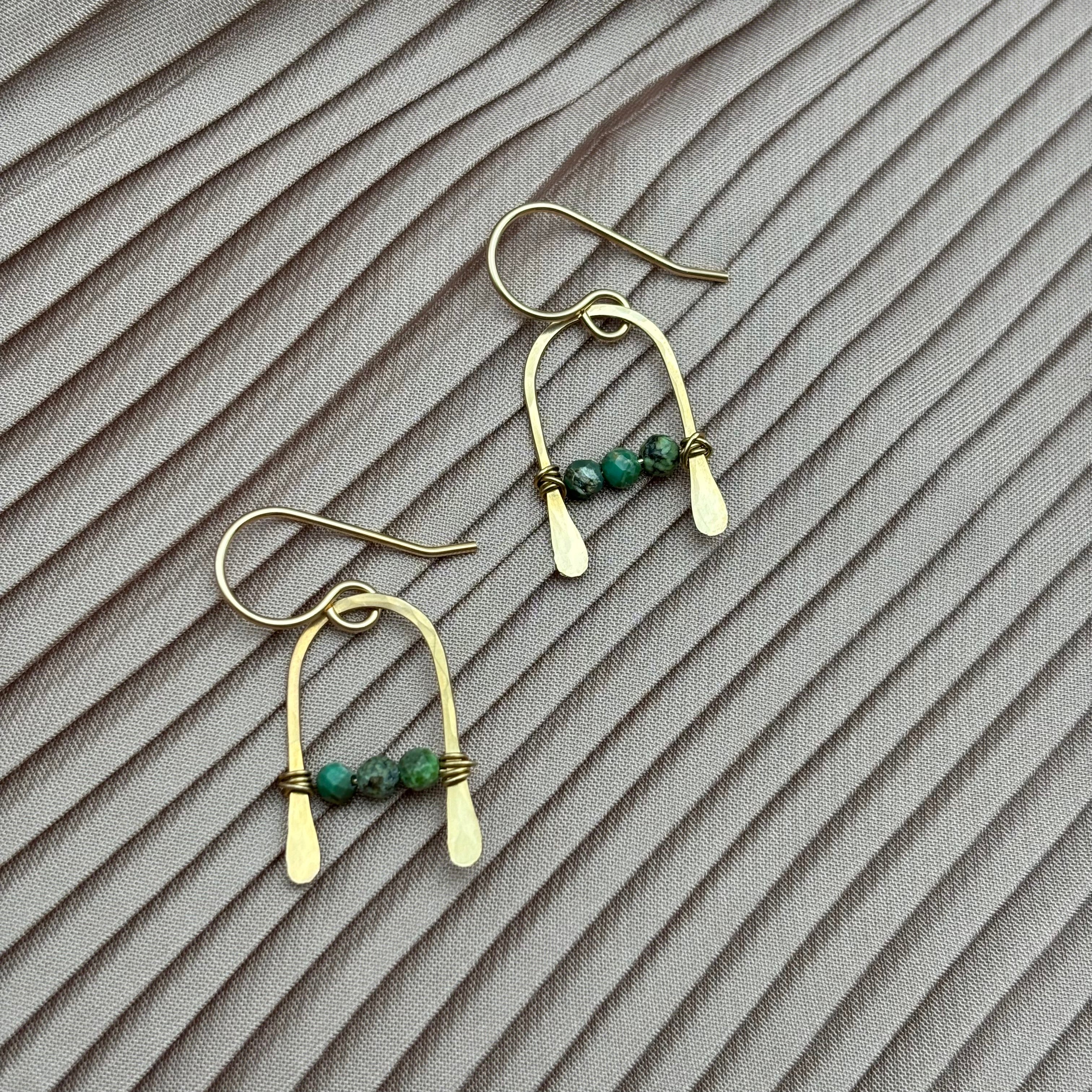 The Bitty Earrings by Fluff Hardware feature an arched gold-filled wire design with hammered ends and three turquoise beads on each, shown on pleated taupe fabric.