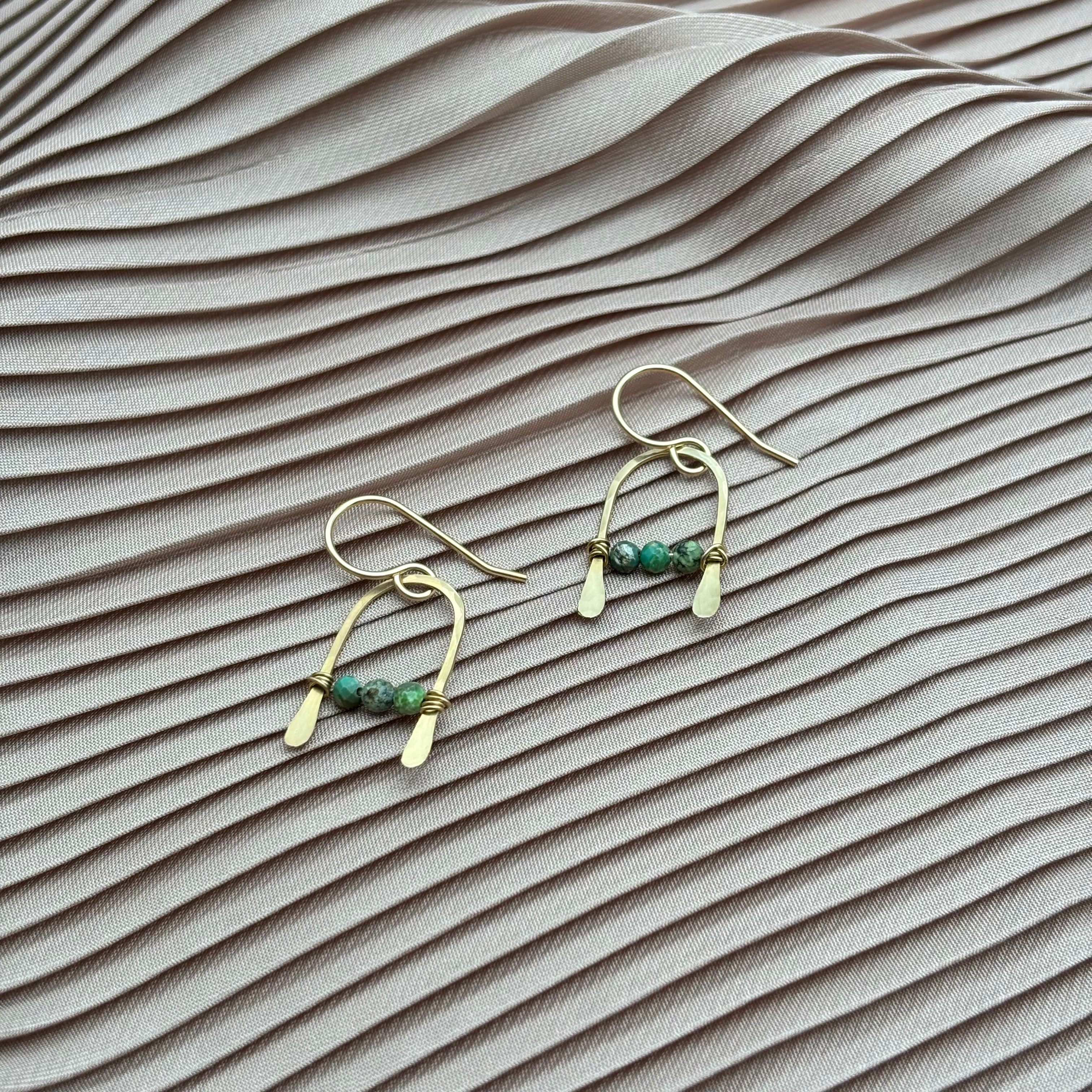 Bitty Earrings by Fluff Hardware, featuring gold filled wire and green beads, are displayed on pale pink pleated satin for a soft, elegant look.