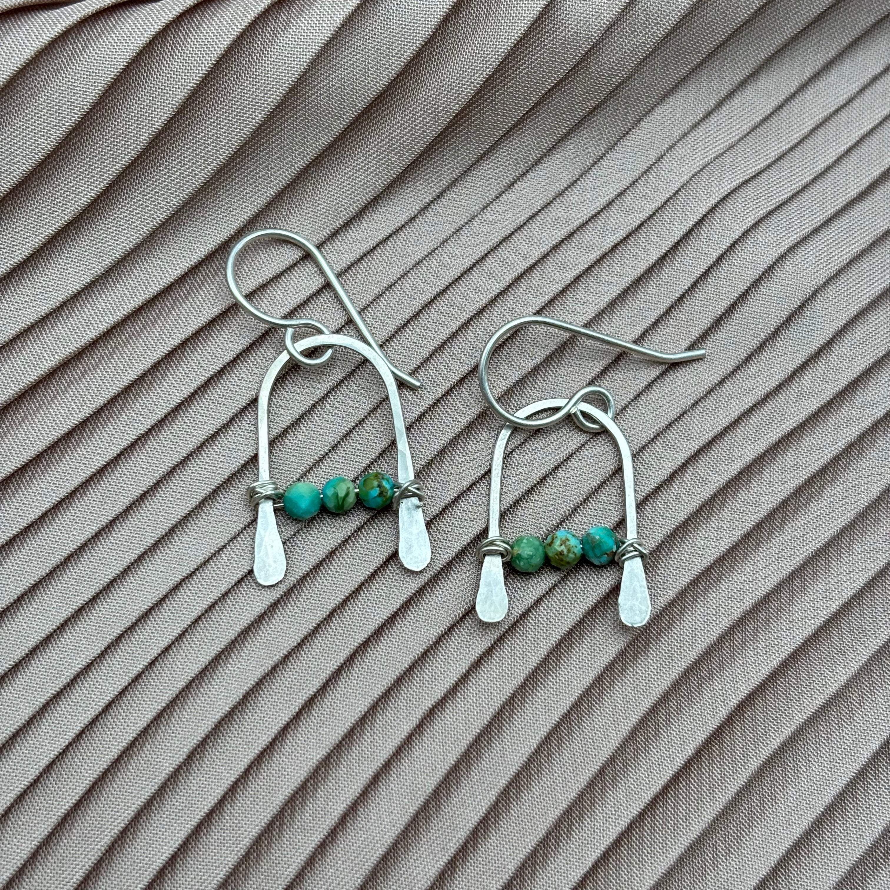 Bitty Earrings by Fluff Hardware: sterling silver dangle earrings featuring three small green and turquoise beads each, displayed on light pink pleated fabric.