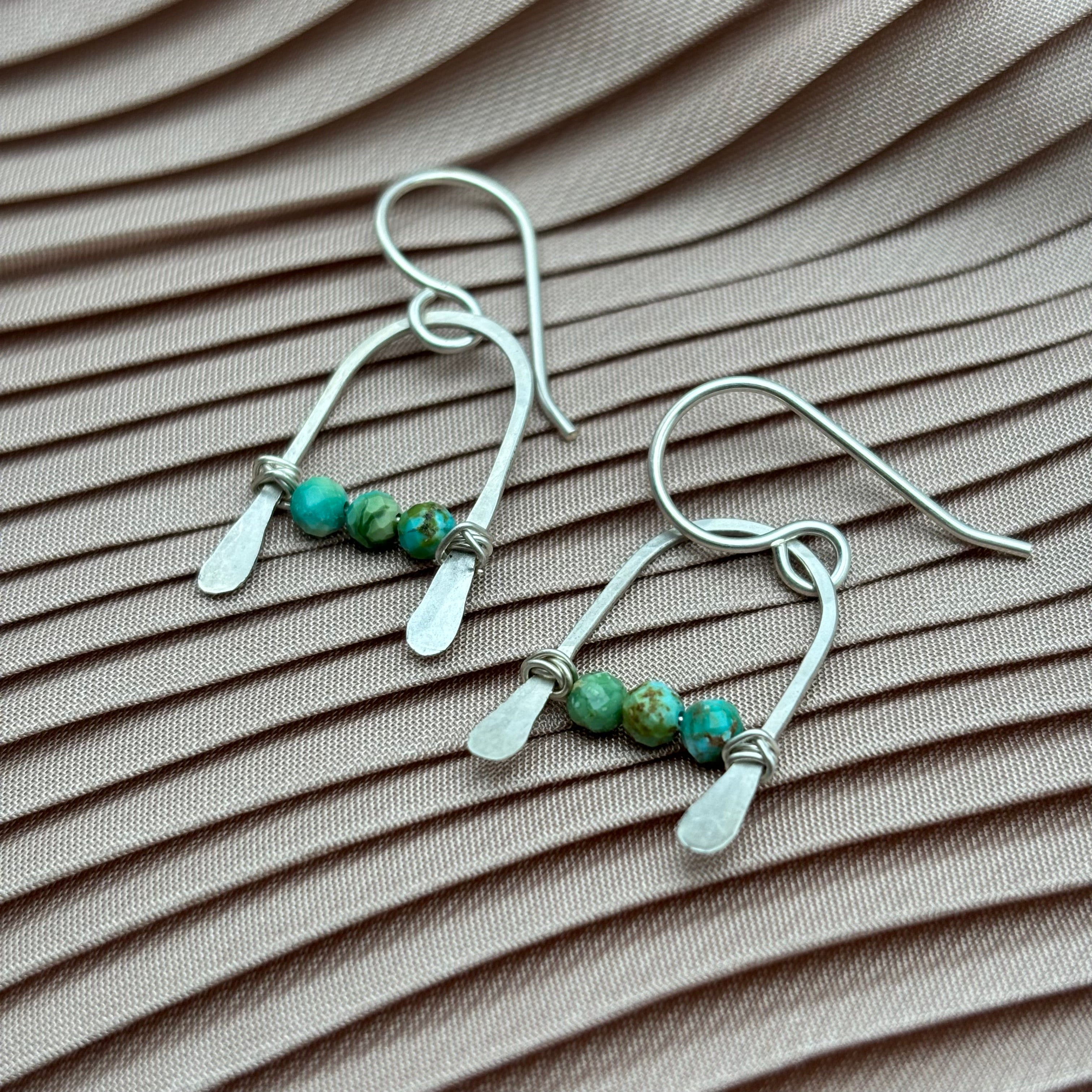 Fluff Hardwares Bitty Earrings feature three turquoise beads on sterling silver wire with hammered accents, shown atop pleated light mauve fabric. The modern, geometric design creates a striking look.